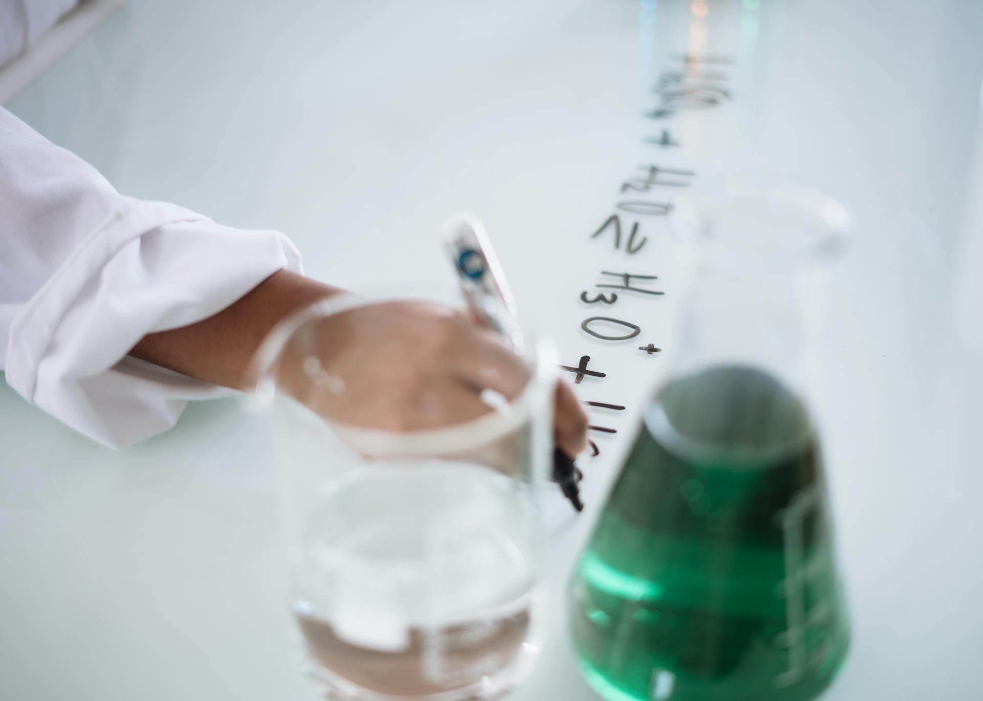 Body part of unrecognizable scientist in white uniform writing down formula after providing chemical research with fluid in flask during science lesson in university