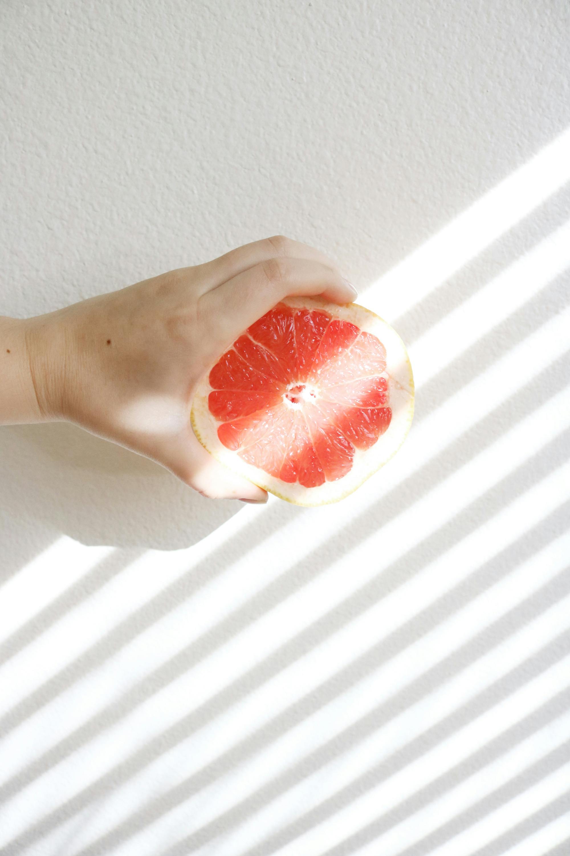 A hand holds a sliced grapefruit against a backdrop of light and shadow stripes.