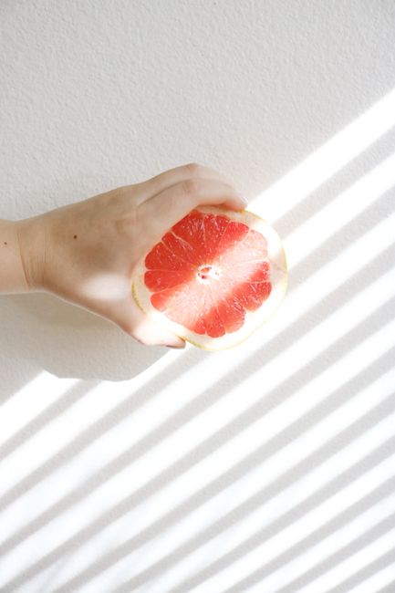 A hand holds a sliced grapefruit against a backdrop of light and shadow stripes.