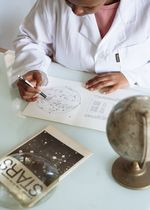 Free Crop of focused scientist wearing white uniform and modifying constellation map while sitting at table with globe and educational materials in university Stock Photo