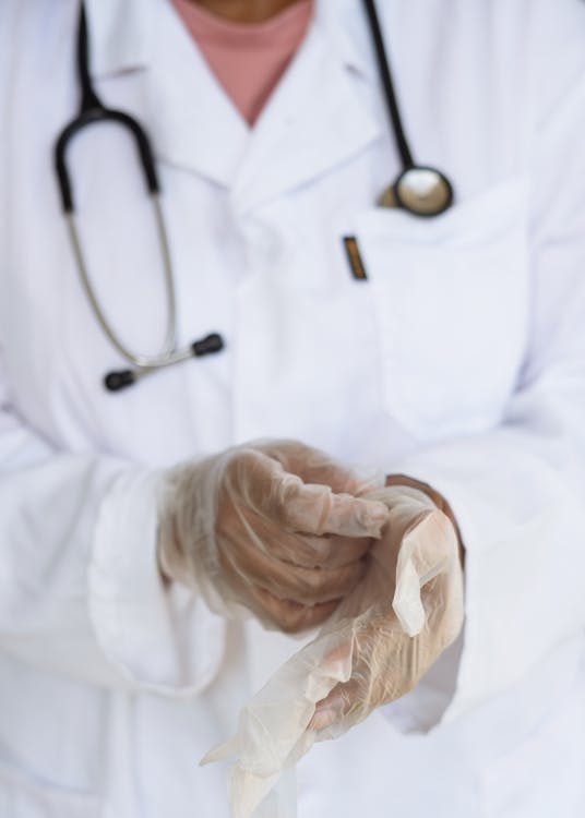 Free Faceless ethnic medical worker in lab coat and stethoscope taking of transparent gloves after approaching patients for examination in modern hospital Stock Photo