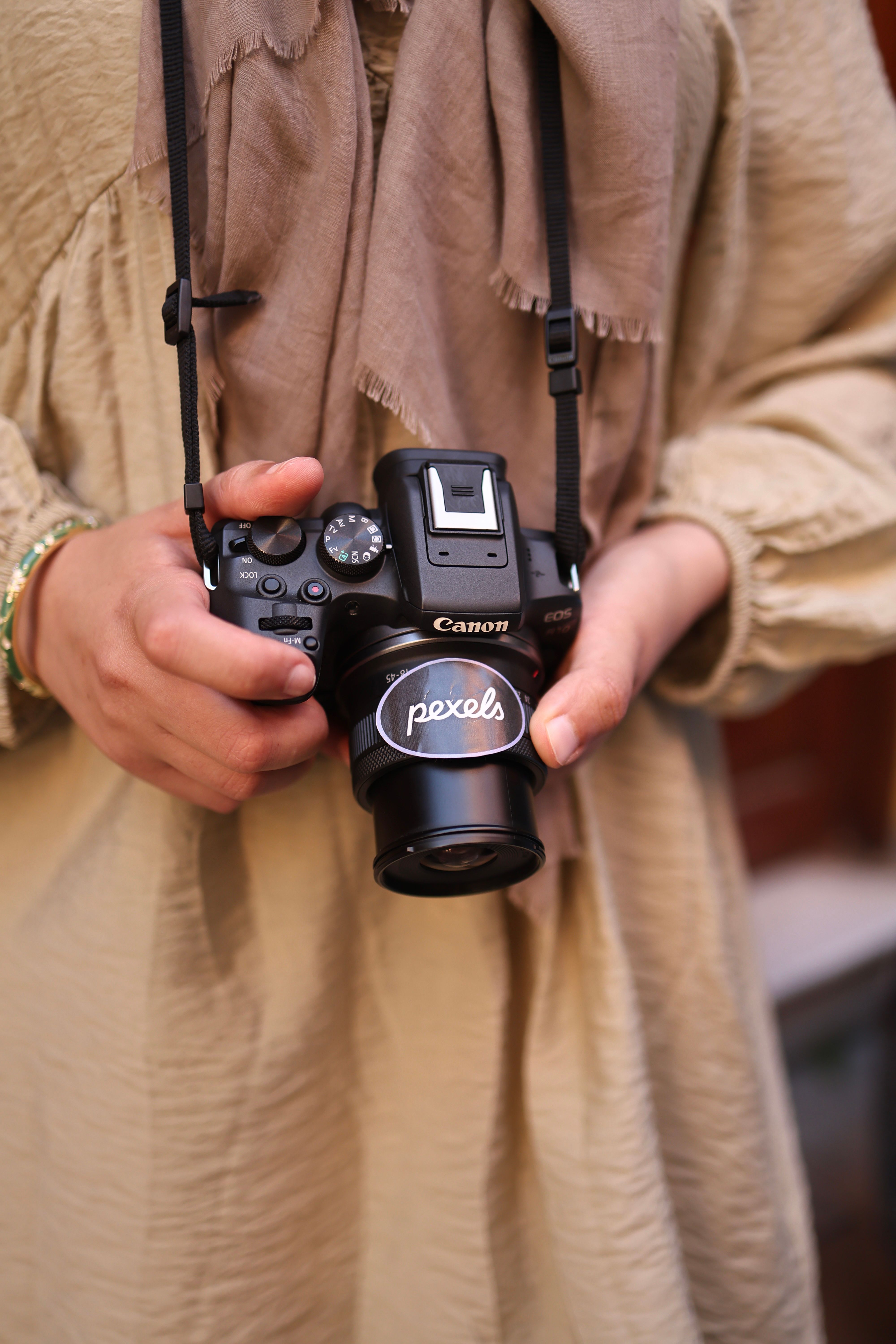 A person gently holds a Canon camera with a lens cap labeled 'Pexels'.