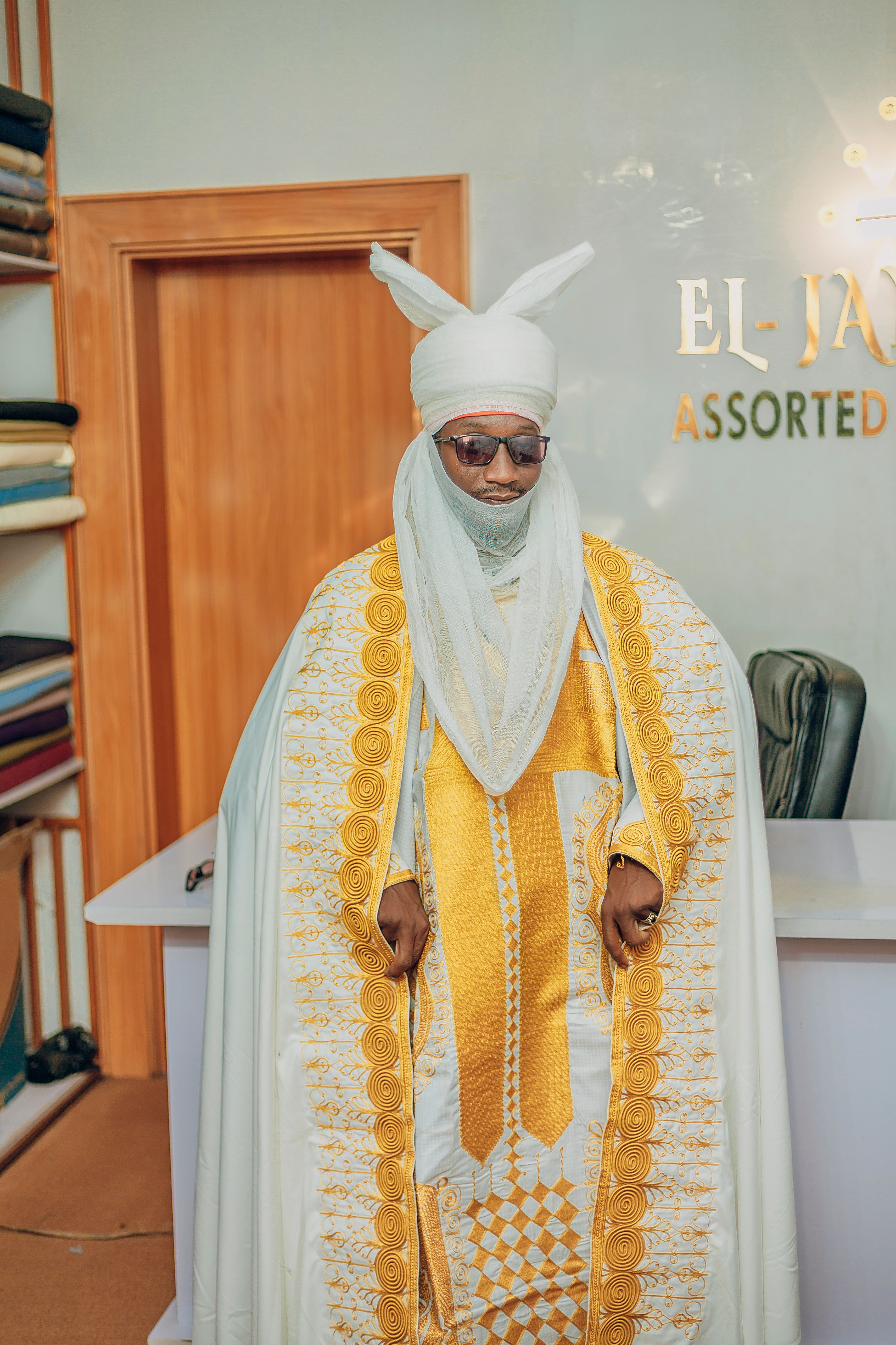 A dignified man in elaborate white and gold traditional clothing inside a room.