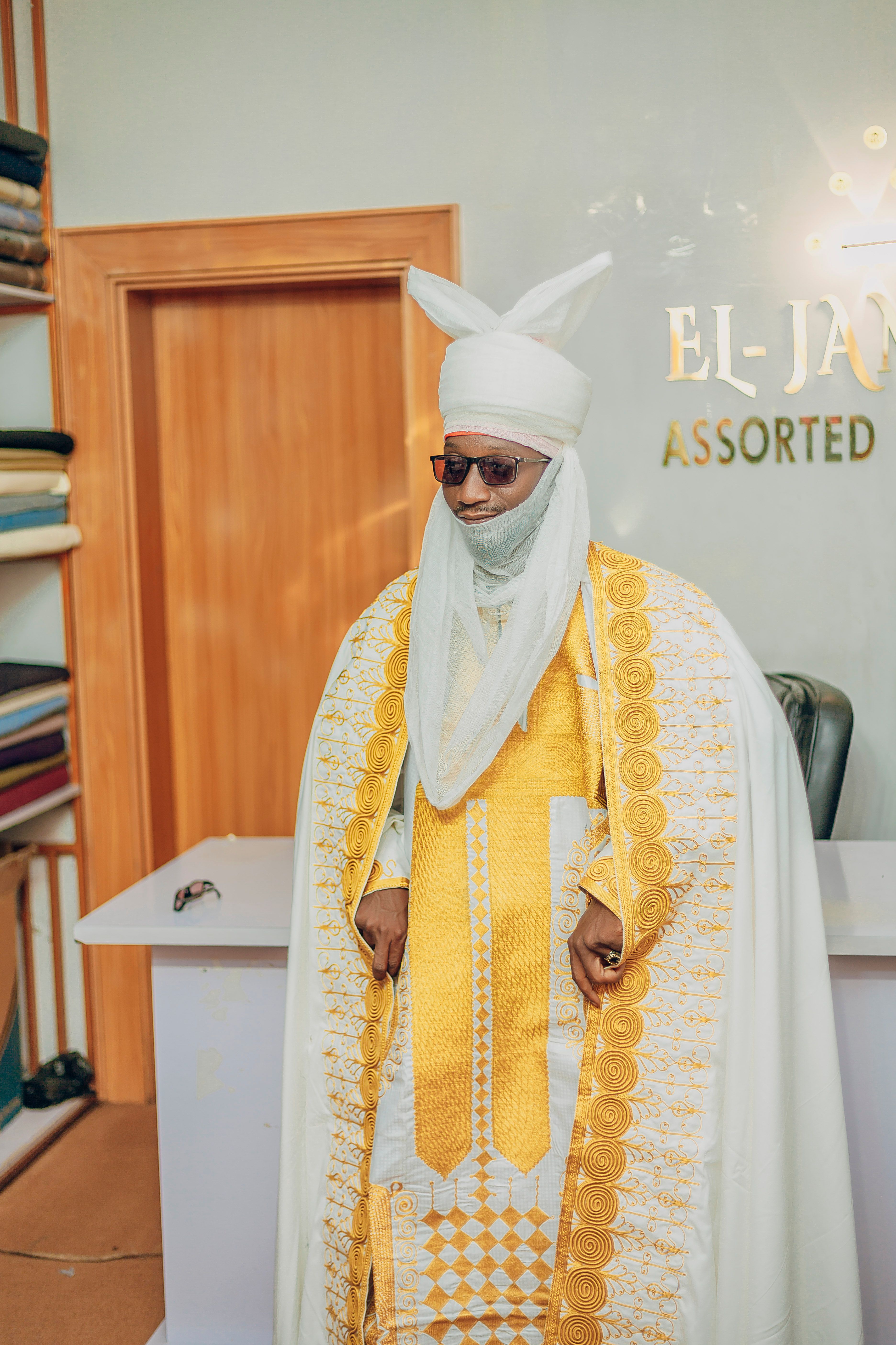 A man in traditional African attire with ornate gold patterns stands indoors by a desk.