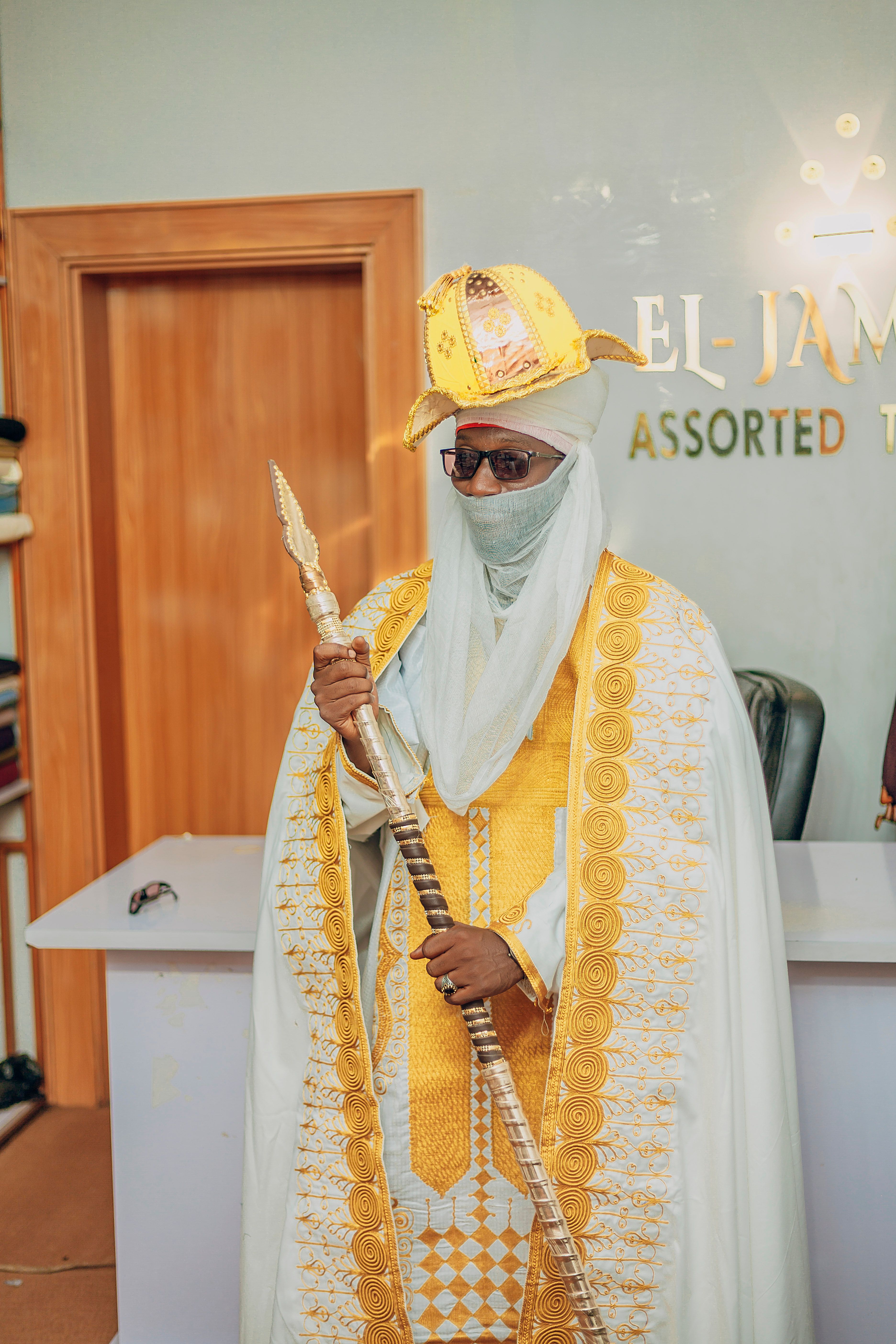 Individual in ornate traditional attire with staff, indoors at El-Jam office.