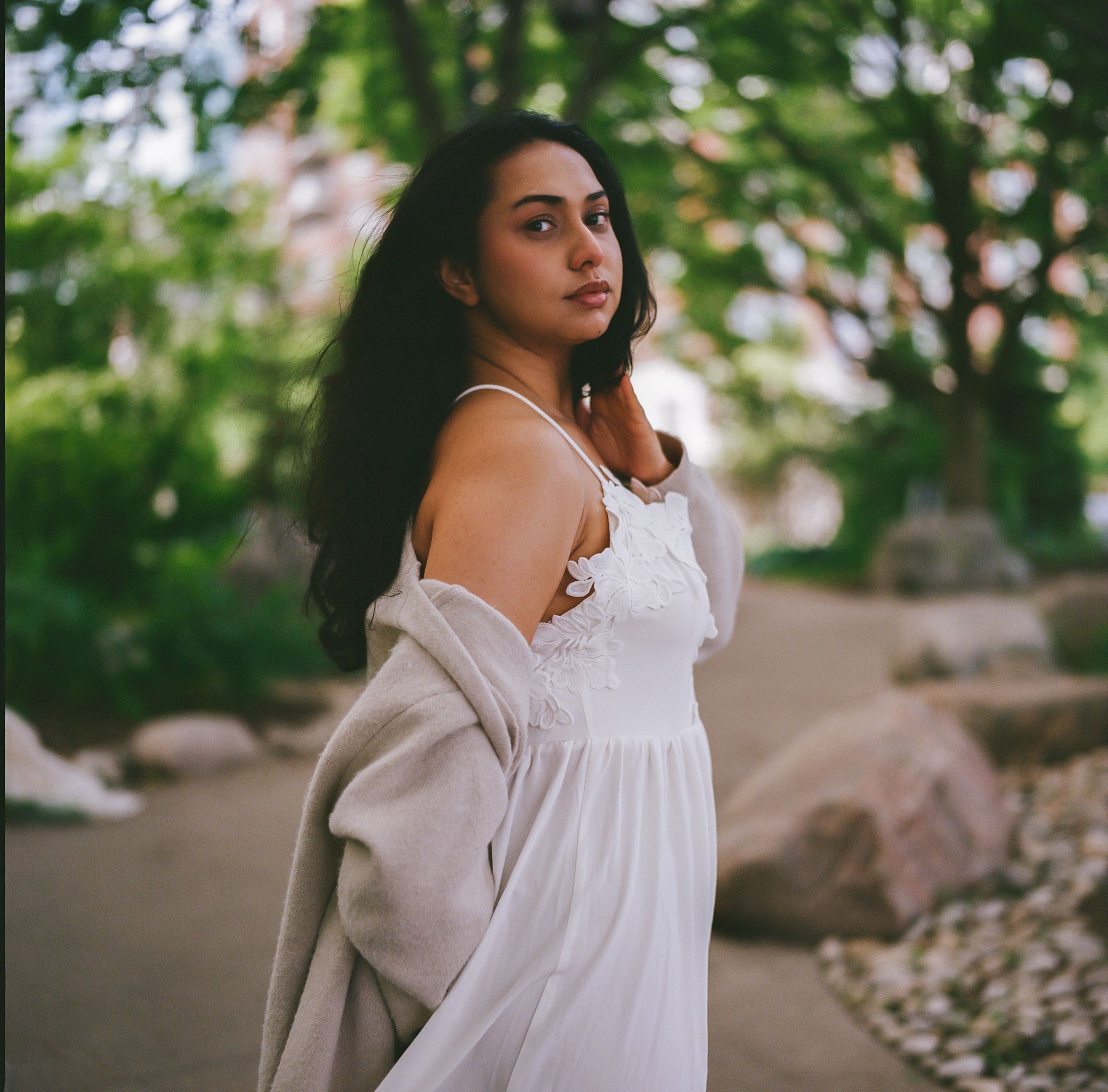 A beautiful woman in a white dress poses elegantly outdoors in a serene park setting.