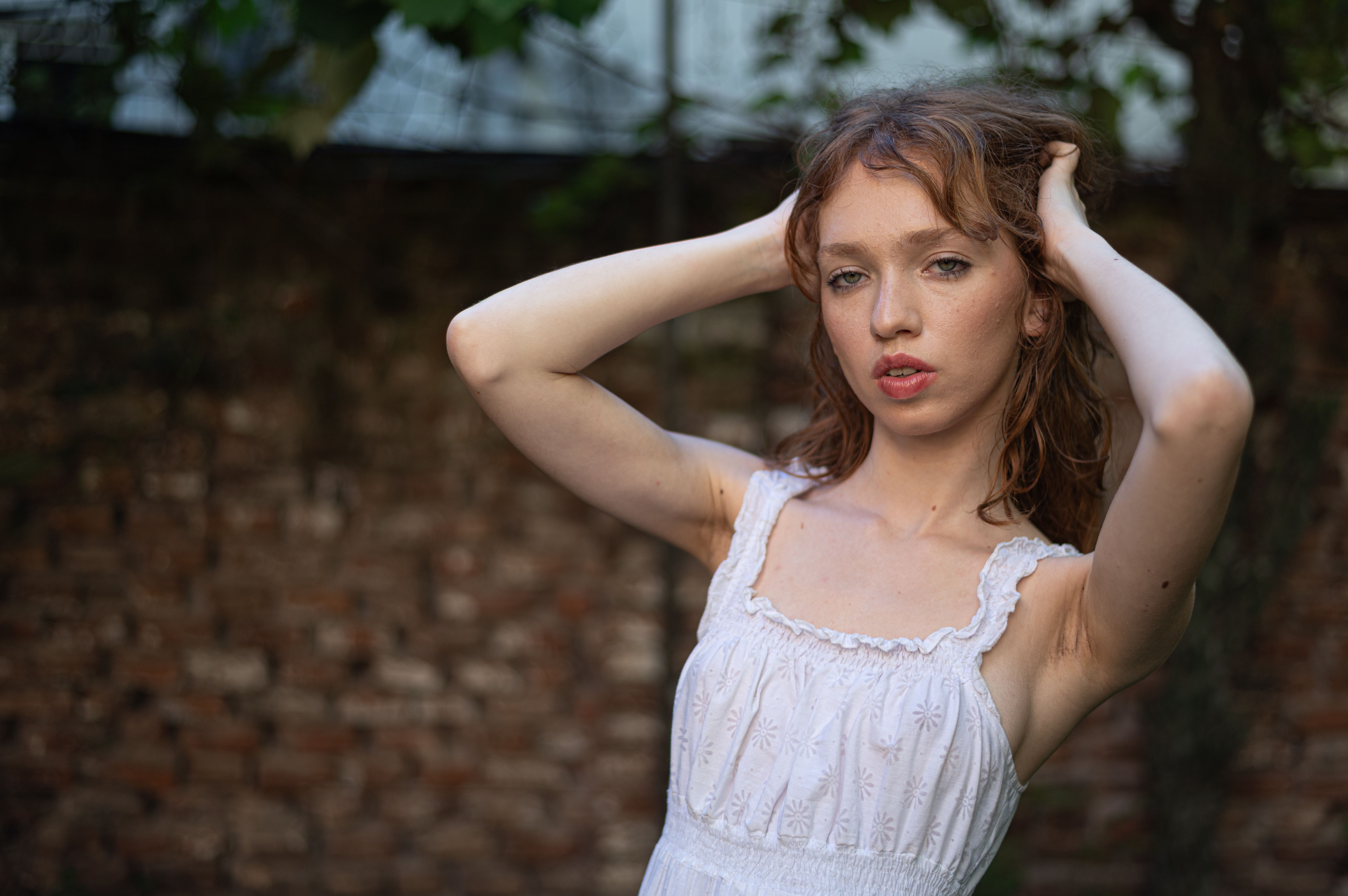 A young woman with red hair poses confidently in a white dress against a brick wall backdrop.