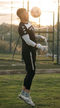 A goalkeeper practicing soccer skills on a grassy field at sunset, wearing full gear and gloves.