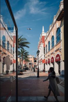 Scenic view of an elegant shopping street with arches, palm trees, and an aircraft overhead.