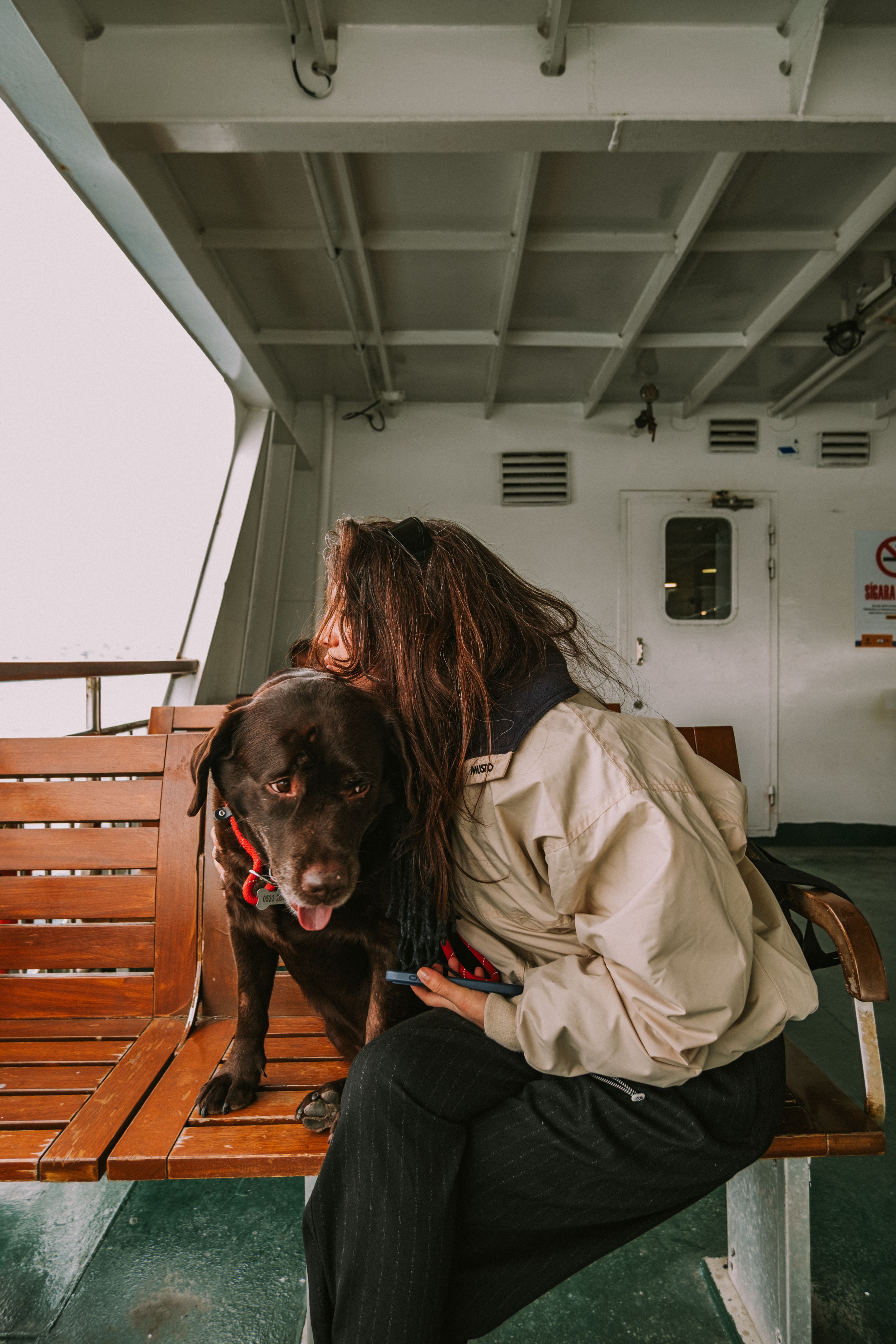 A woman in a coat sits with her Labrador dog on a ferry bench, creating a warm and cozy moment.