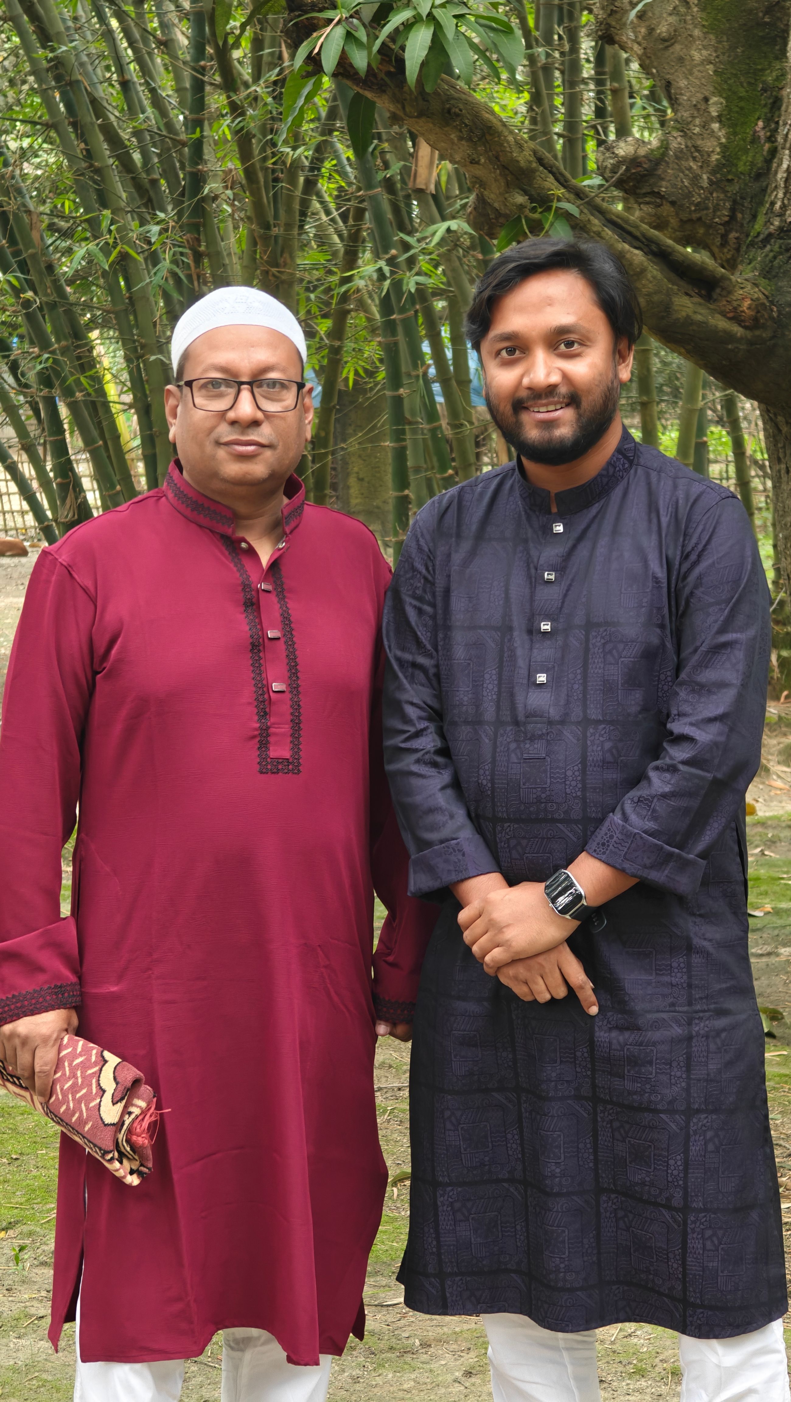 Two men standing together in traditional South Asian clothing outdoors with a bamboo background.