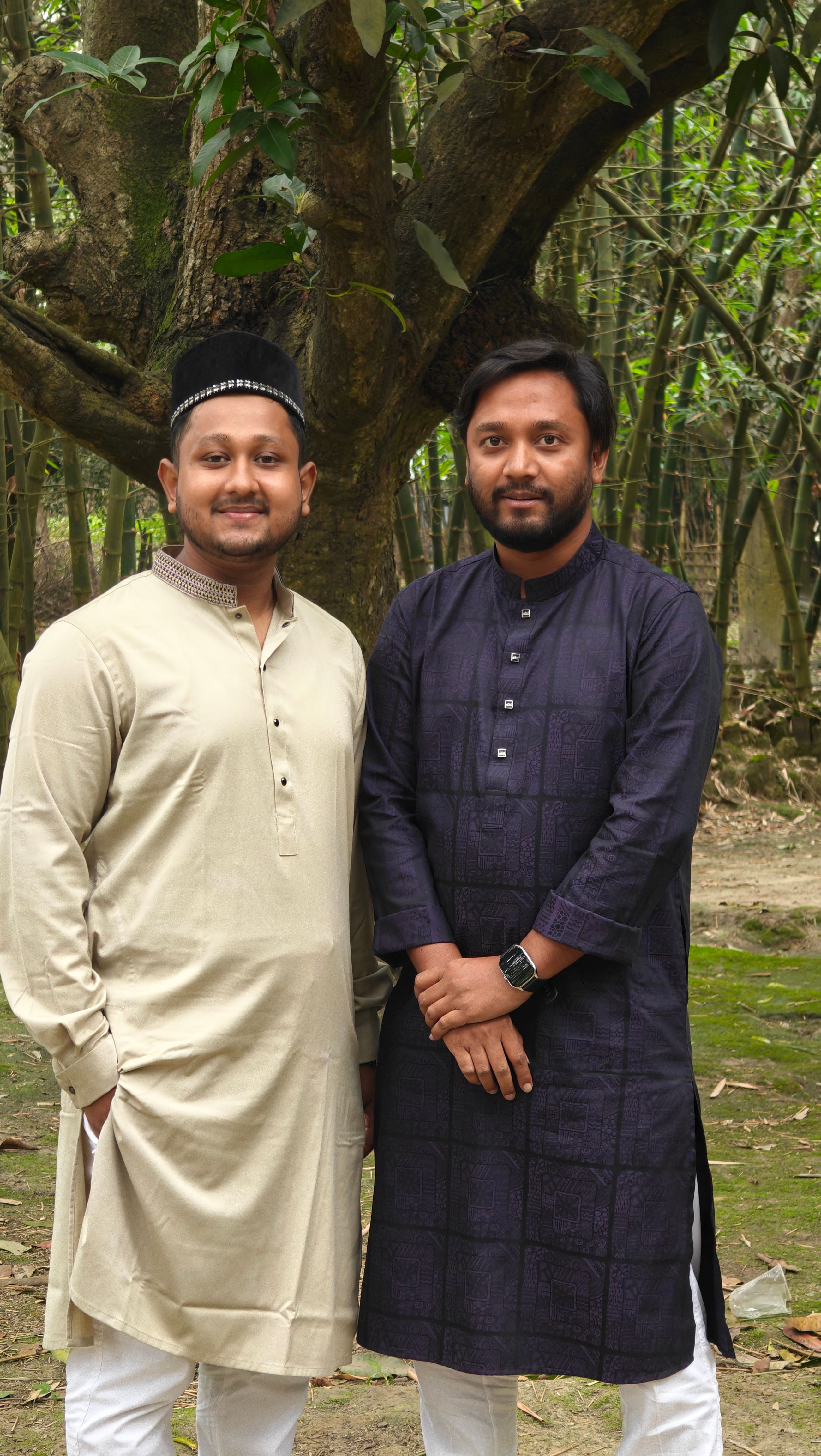 Two men in traditional clothing posing outdoors with trees in the background.
