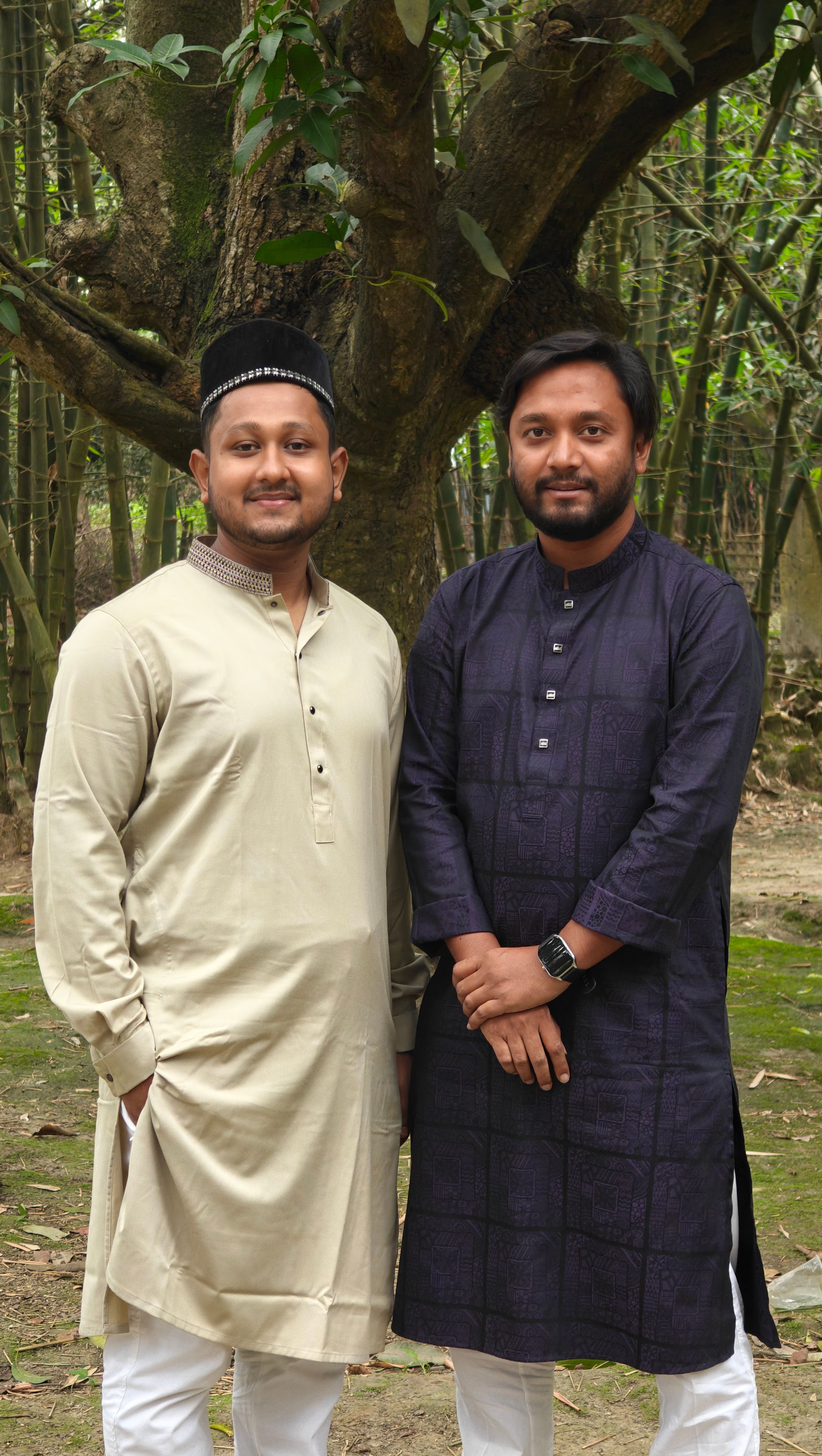 Two men wearing traditional clothing pose outdoors under a tree.