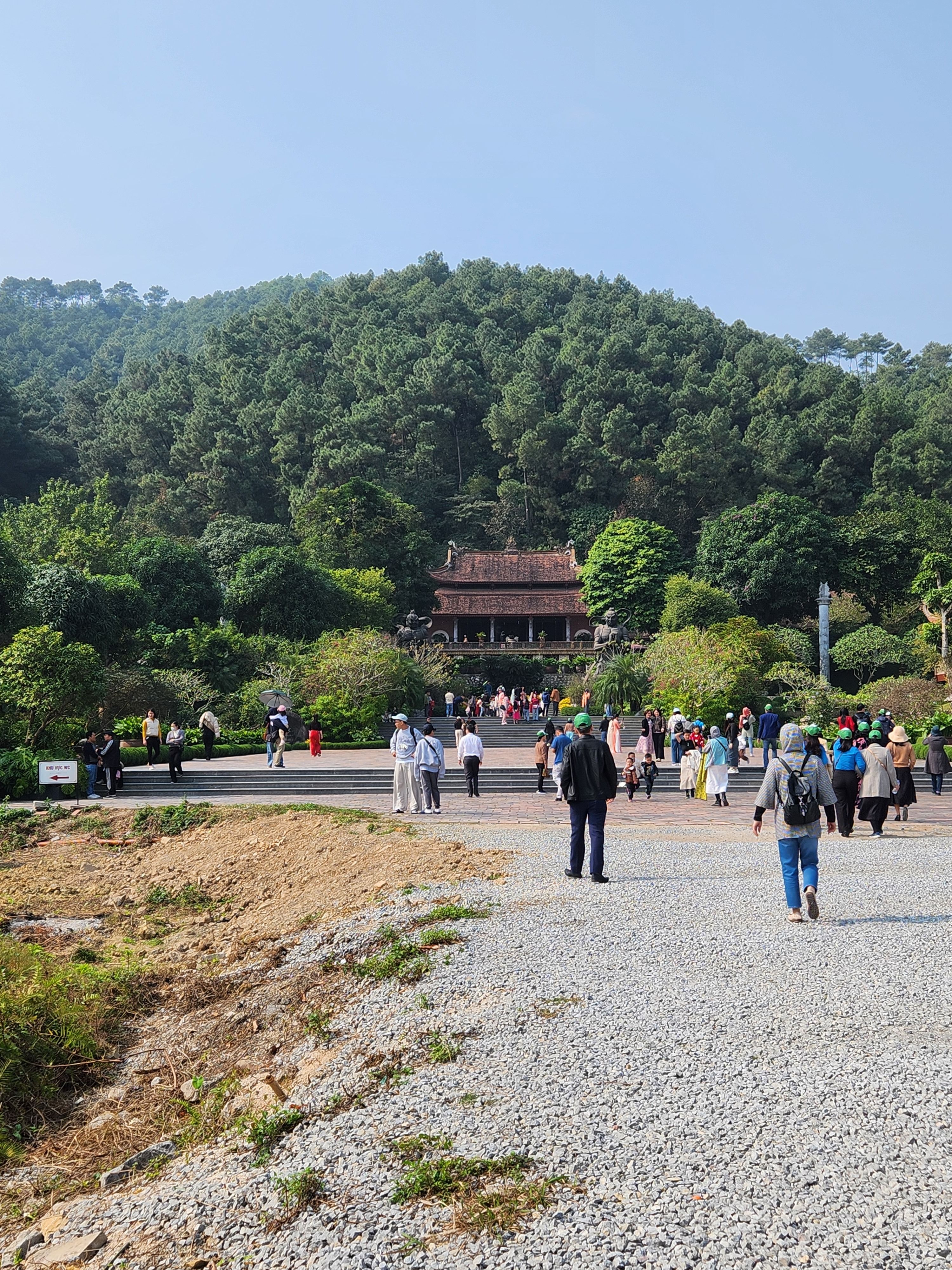 Visitors explore a Vietnamese temple set against lush hills under a clear sky, showcasing cultural architecture.