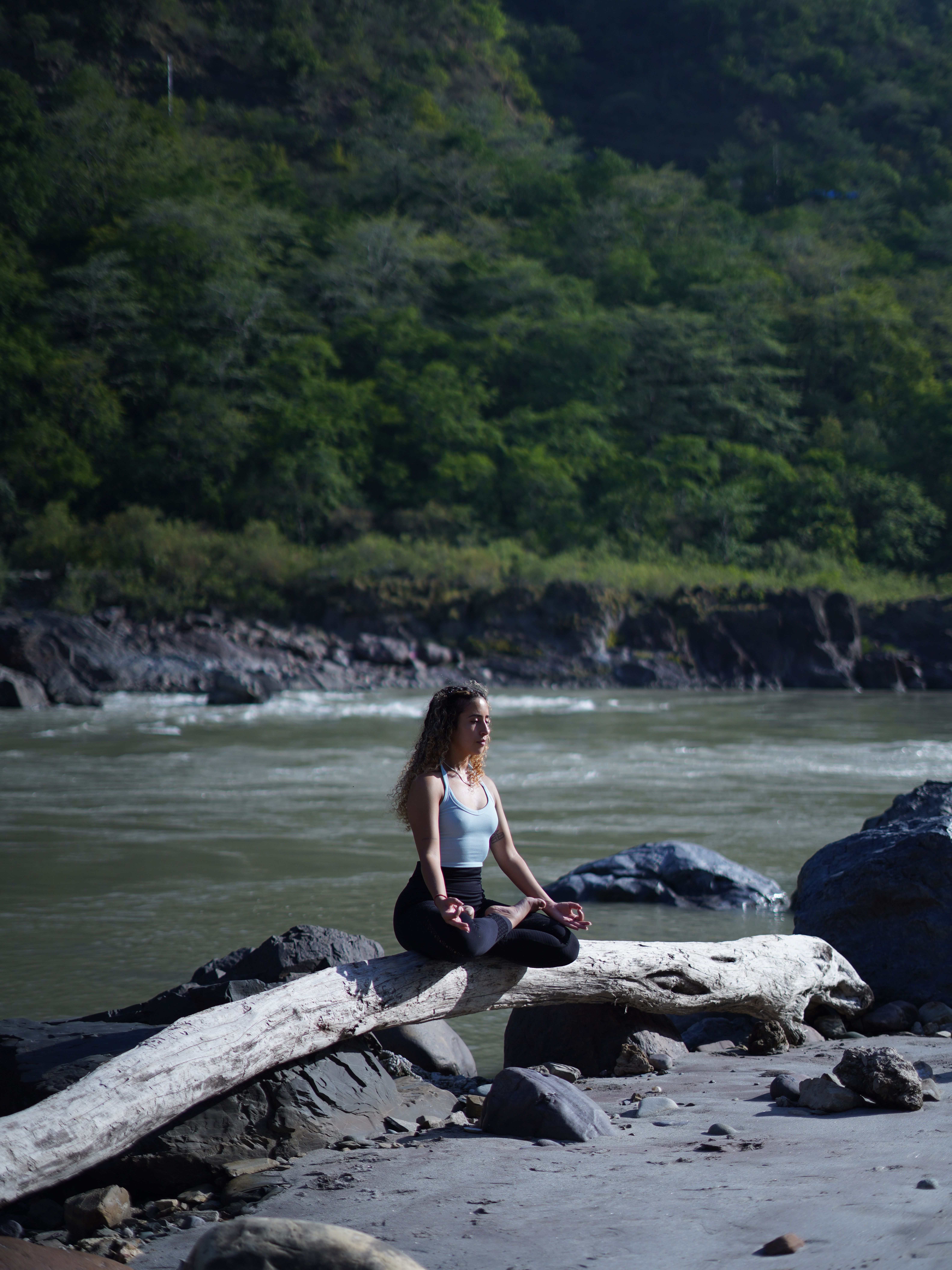 A woman practices meditation on a log by a riverside, surrounded by lush forests.