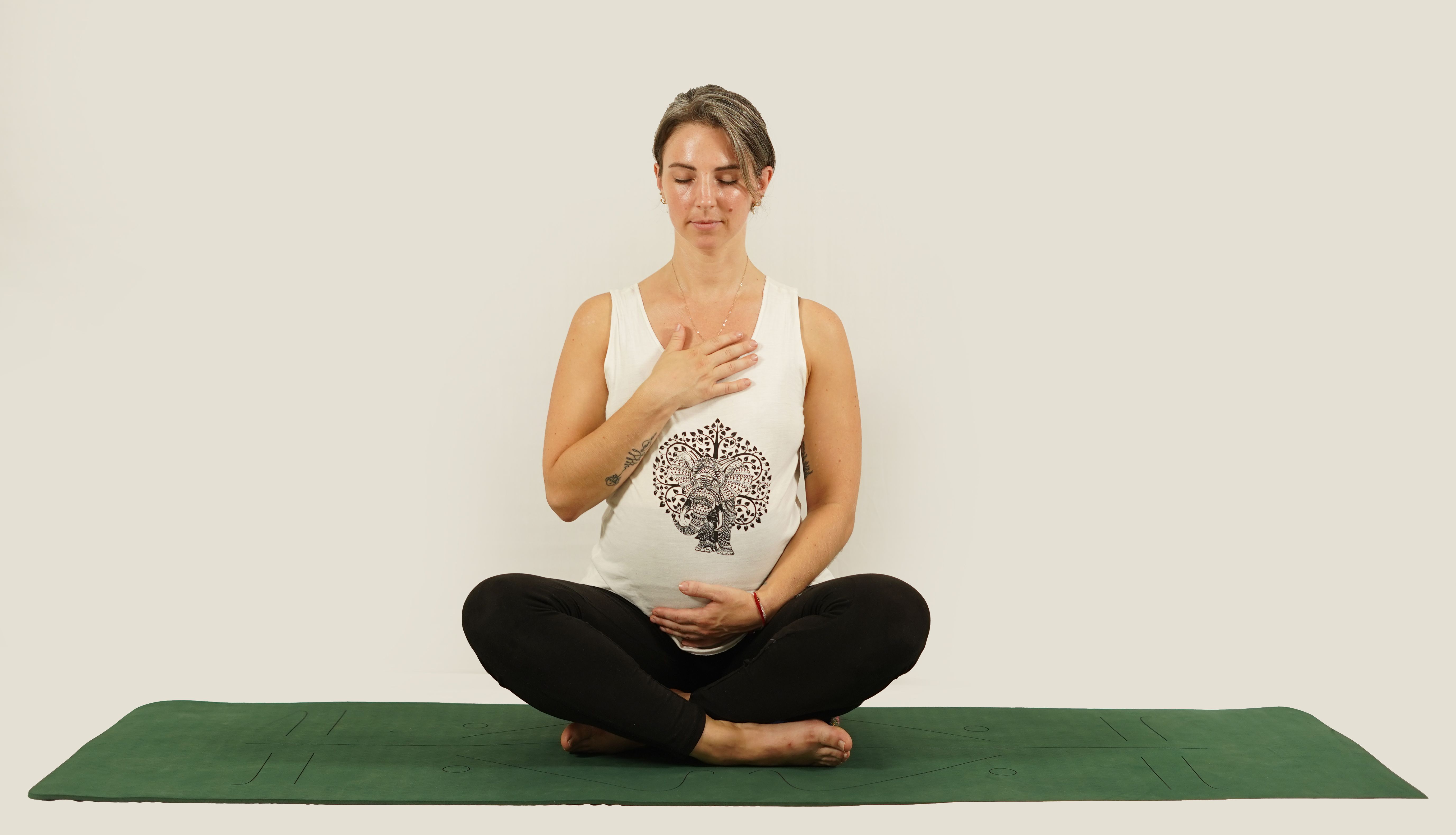Pregnant woman sitting cross-legged on a green yoga mat, meditating with a calm expression indoors.