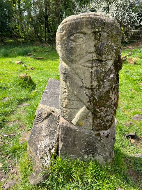 Fotomural Antigua escultura de piedra cubierta de musgo, rodeada de exuberante hierba verde en un apacible entorno rural irlandés.