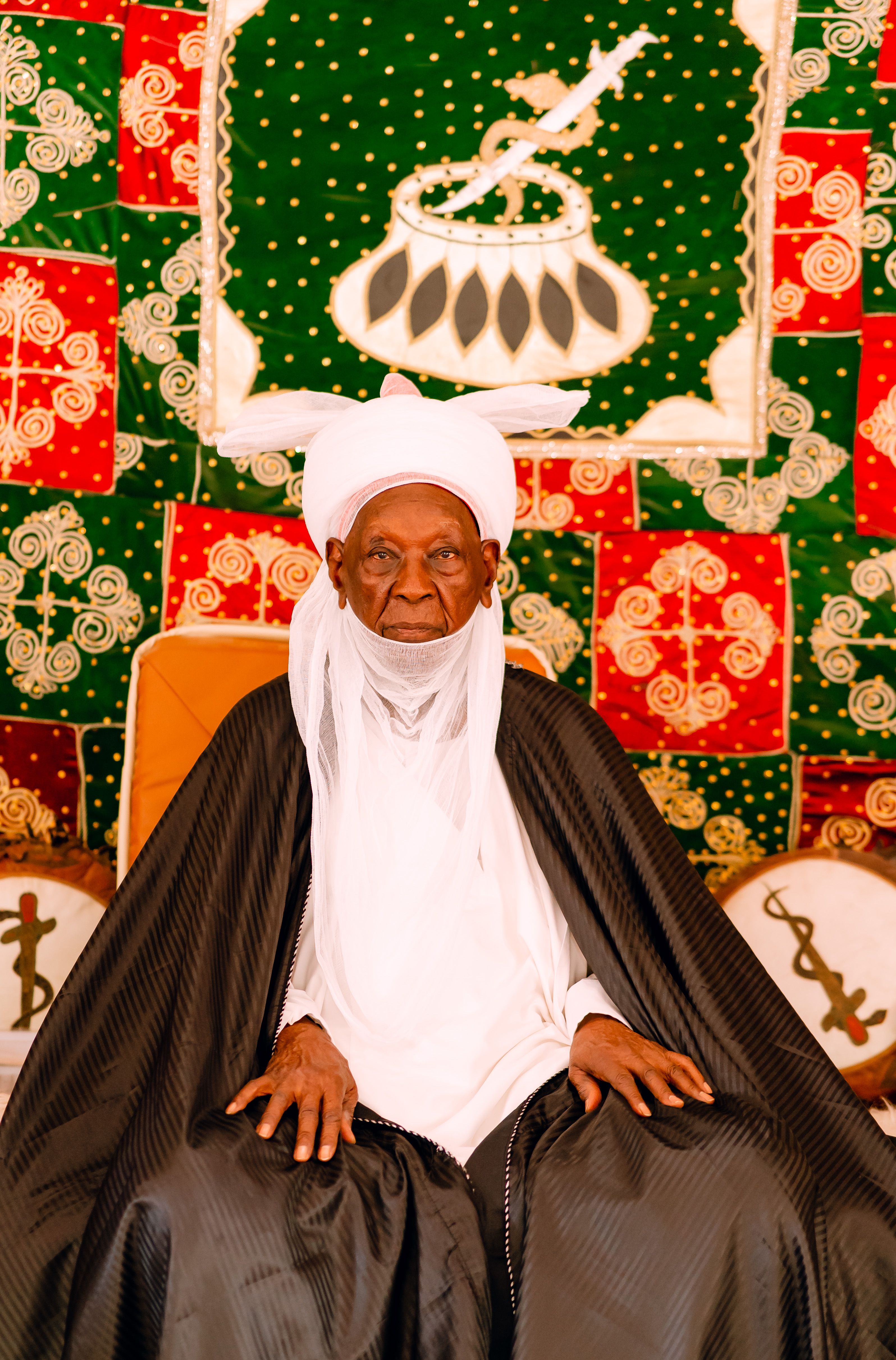 Elderly man in traditional clothing sitting against a vibrant patterned background.