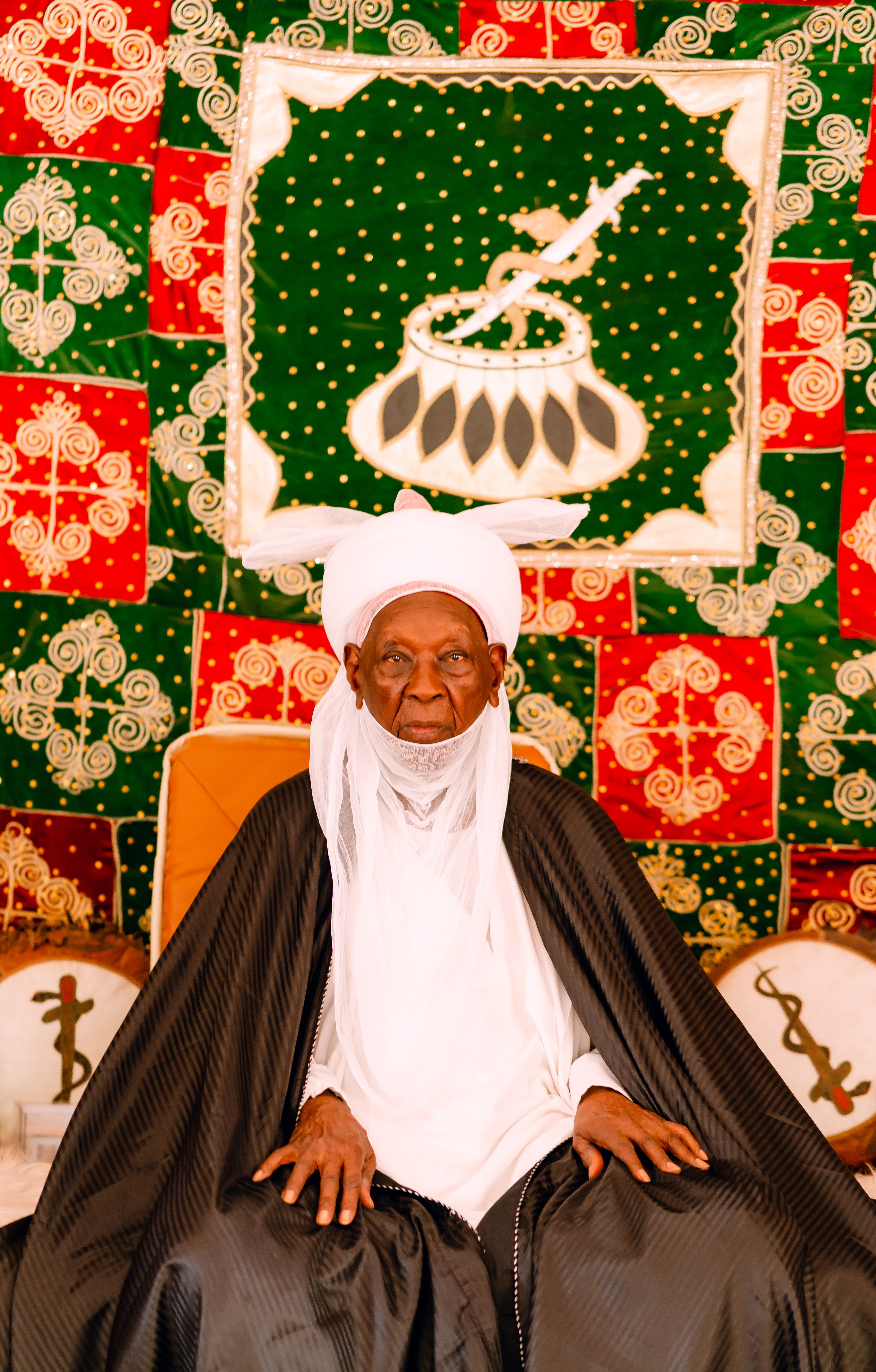 Elderly man in vibrant traditional African attire seated in front of colorful textile backdrop.