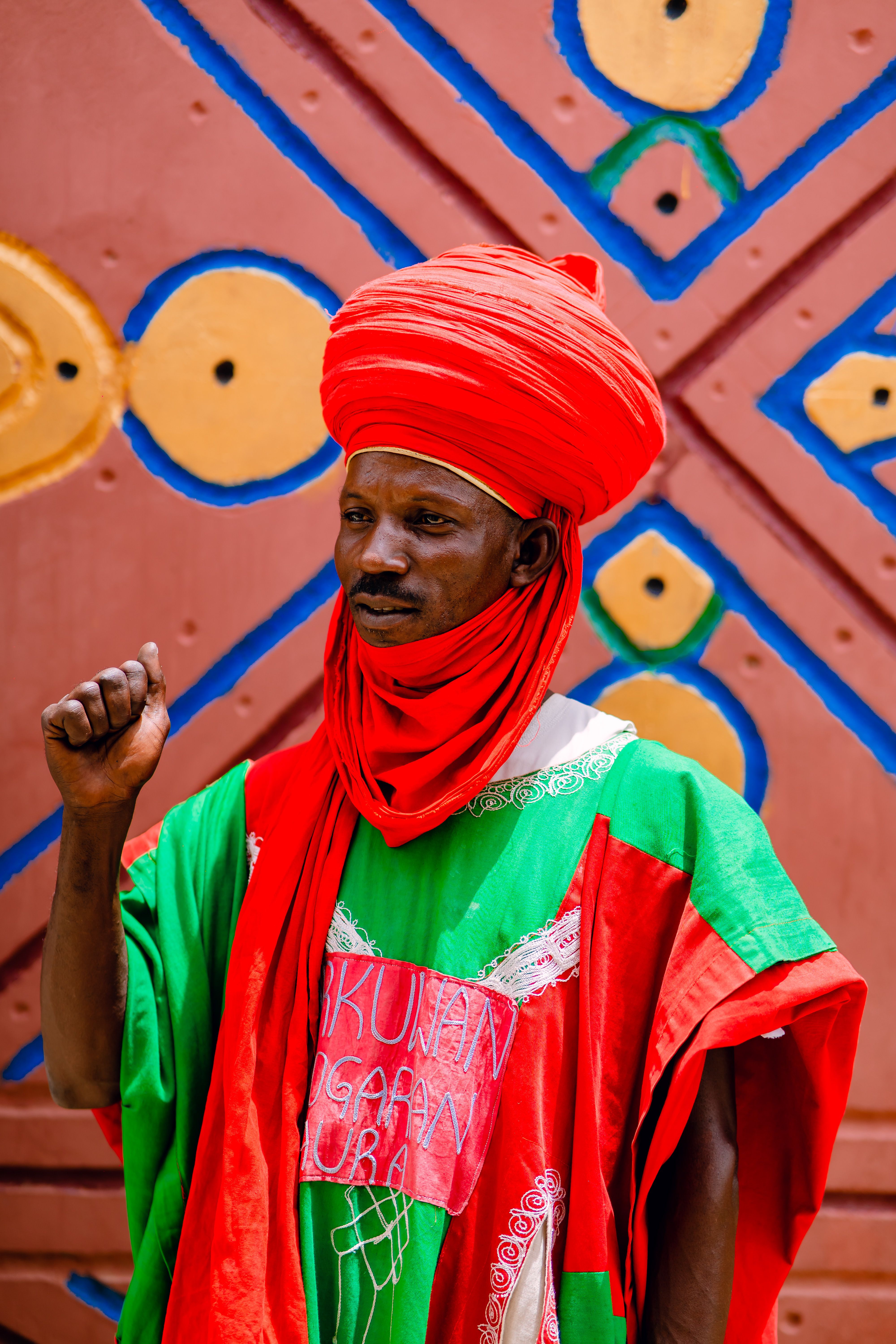 Man in vibrant traditional attire with intricate background pattern.