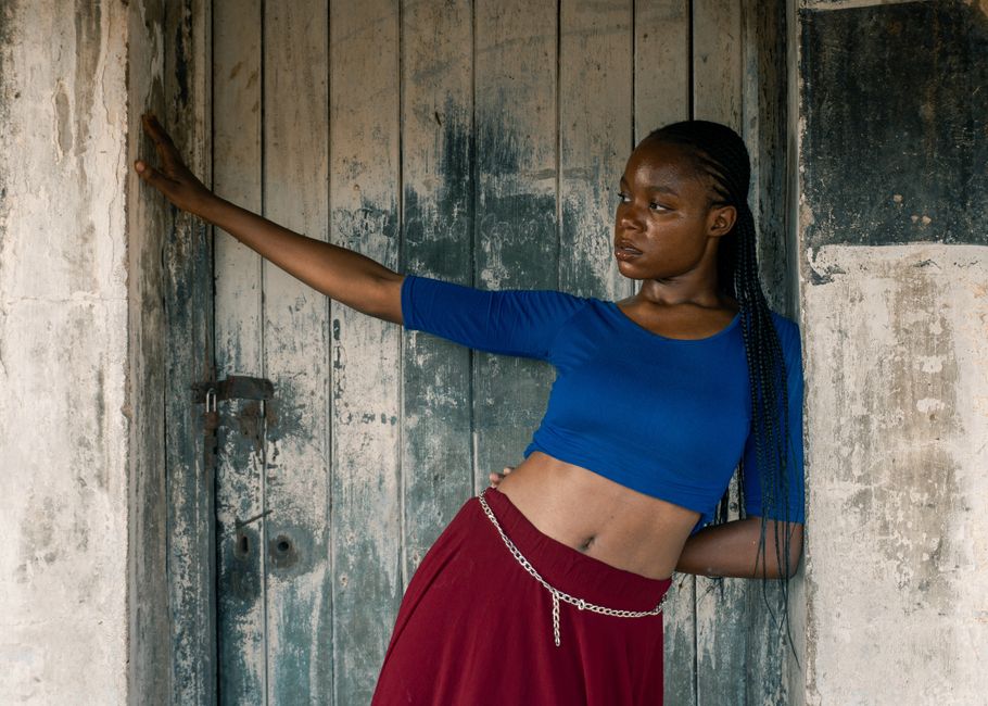 A poised woman poses against a weathered wooden door, wearing a blue crop top and red skirt, exuding confidence.
