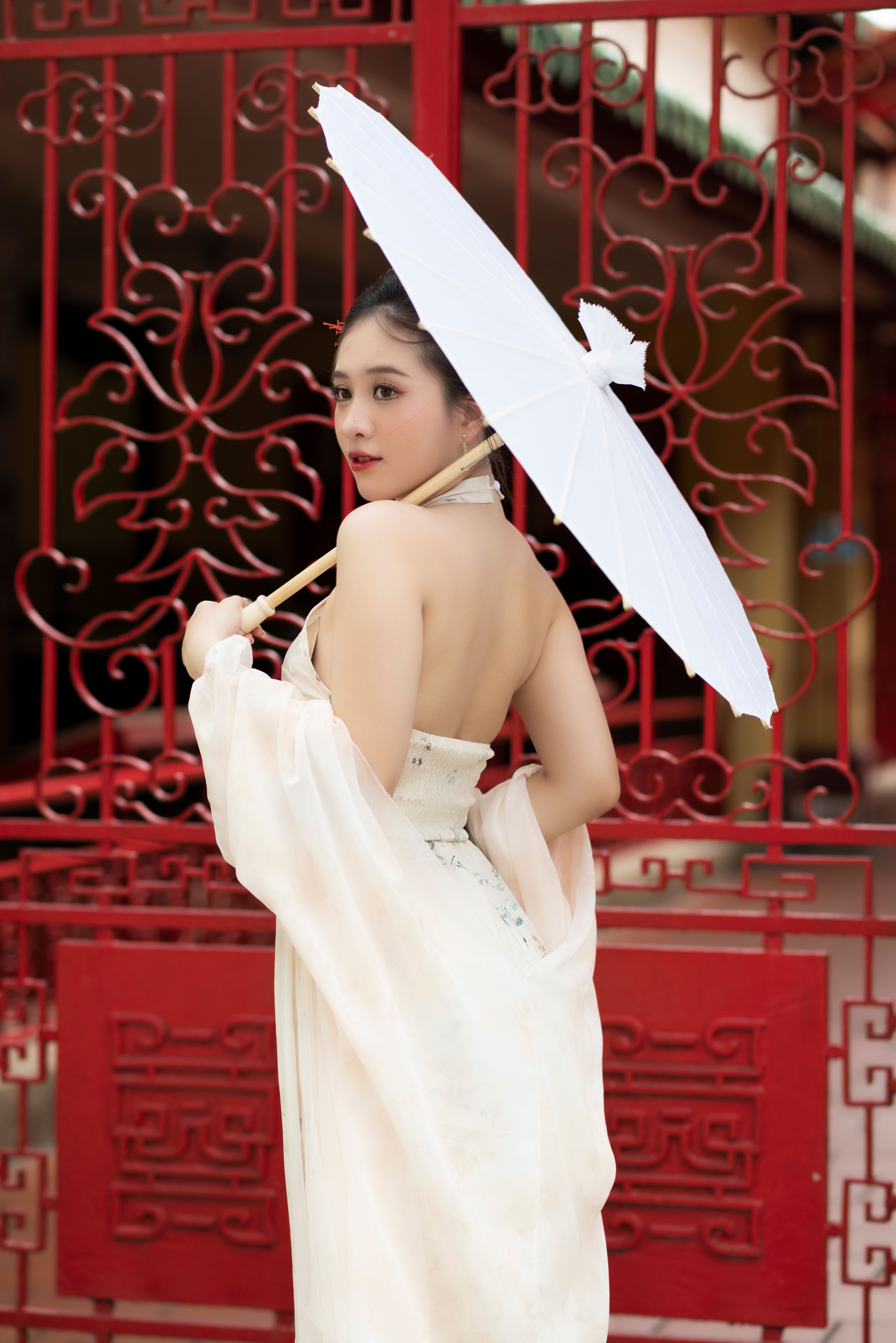 An elegant woman in traditional attire poses with a white umbrella in front of an ornate red gate.