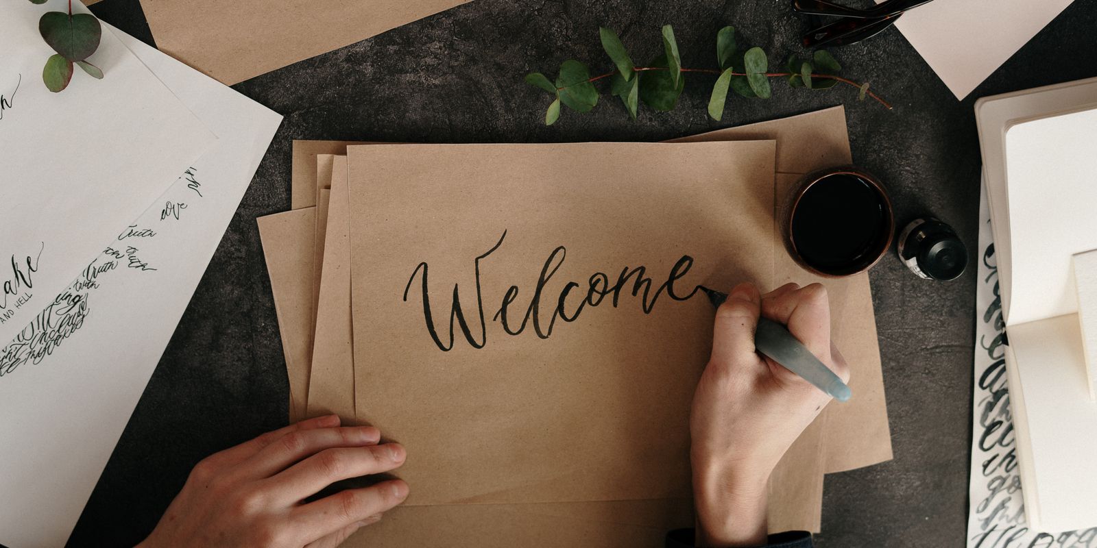 A hand lettering the word "Welcome" in ink calligraphy on kraft paper