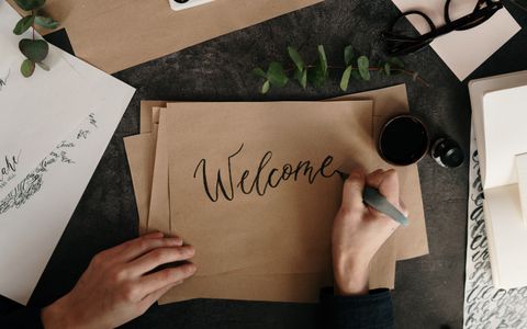 A hand lettering the word "Welcome" in ink calligraphy on kraft paper