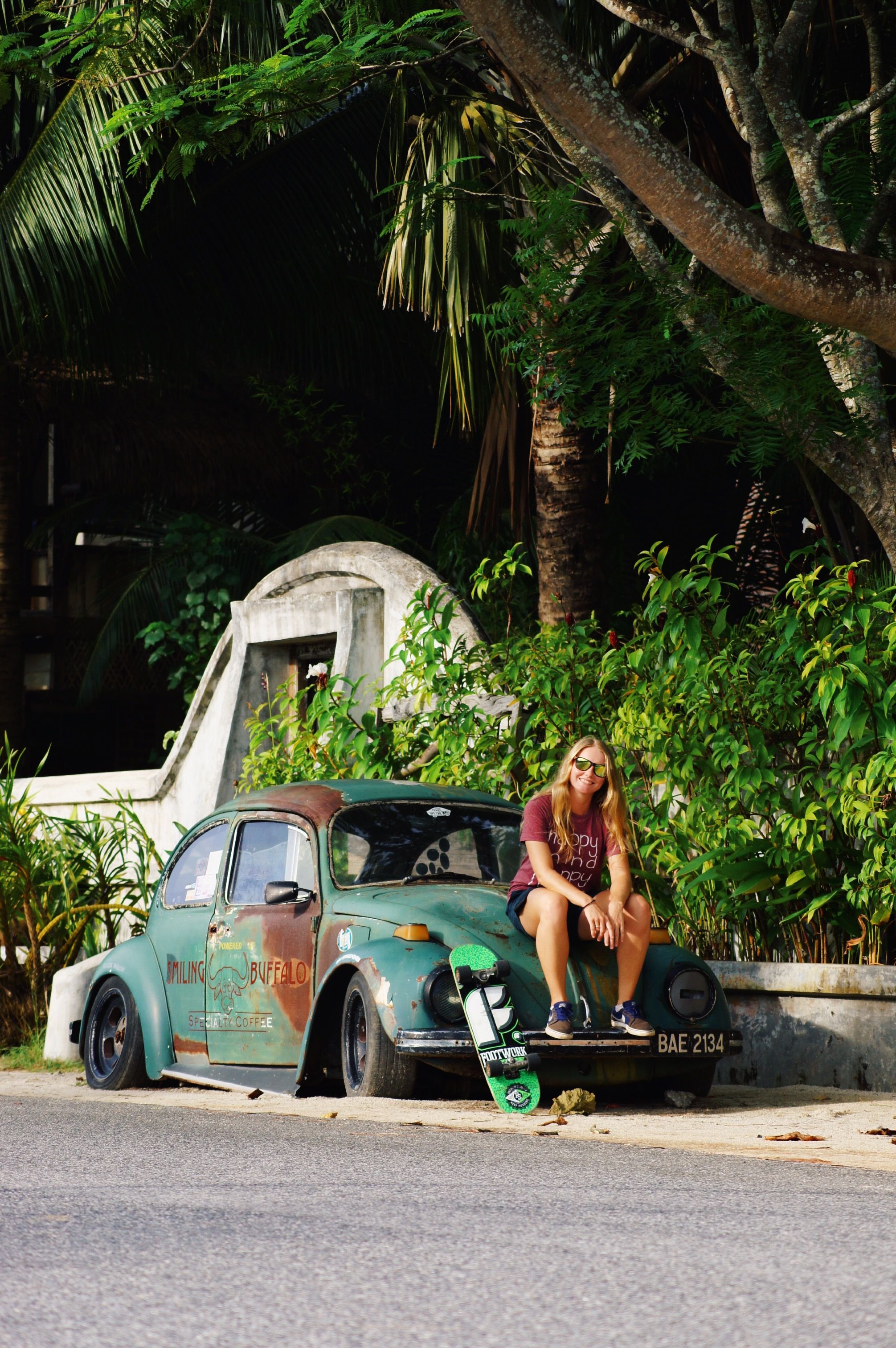 A young woman sits on a classic car with skateboard in lush outdoor setting.