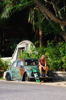 A young woman sits on a classic car with skateboard in lush outdoor setting.