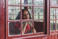 一位沉思的年轻少数族裔女性，留着波浪长发，白天站在窗前，将头靠在拳头上，透过玻璃发呆