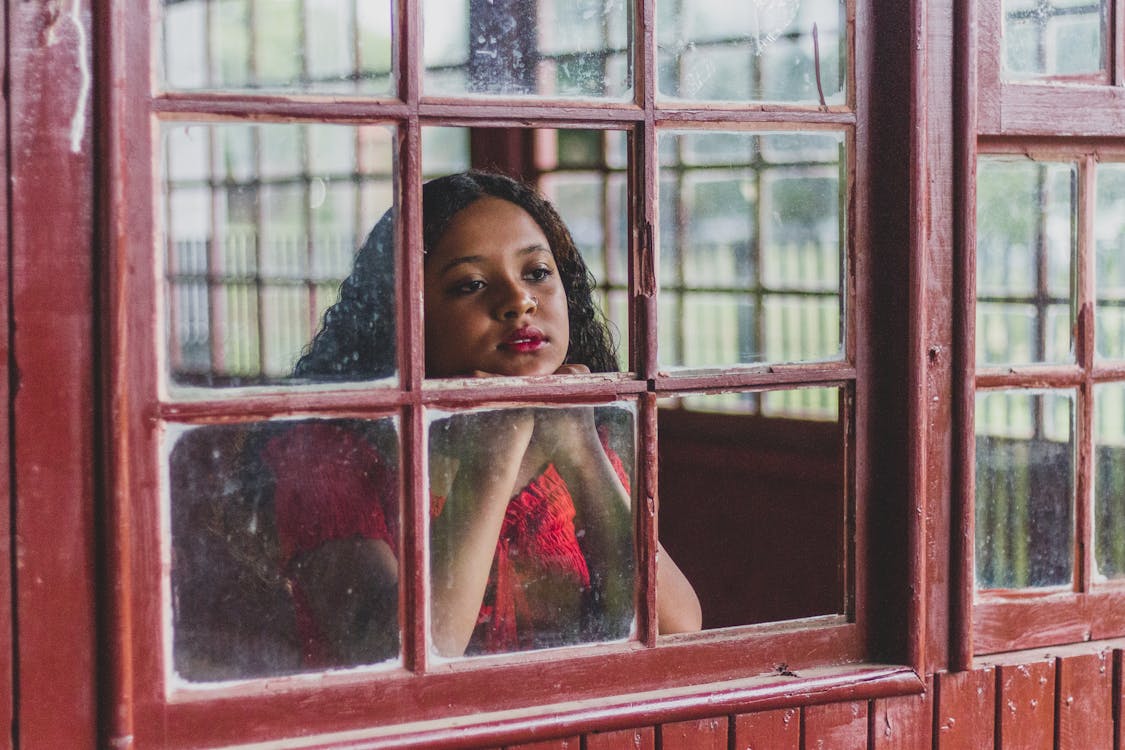 Free Pensive young ethnic female with wavy hair leaning head on fists while standing near window and dreamily looking through glass away in daytime Stock Photo