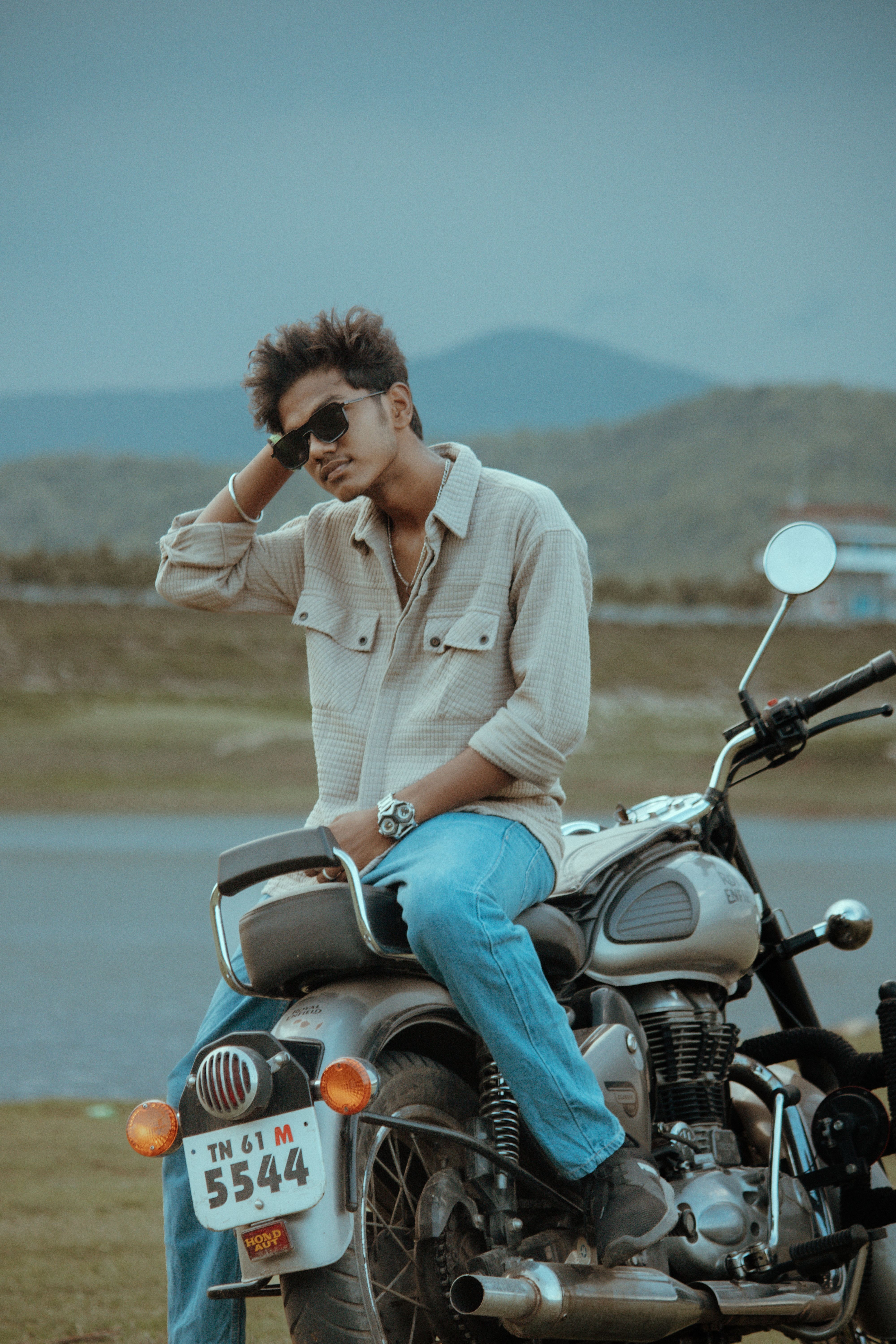 A young man in sunglasses leans on a vintage motorcycle by a lake with mountains in the background.