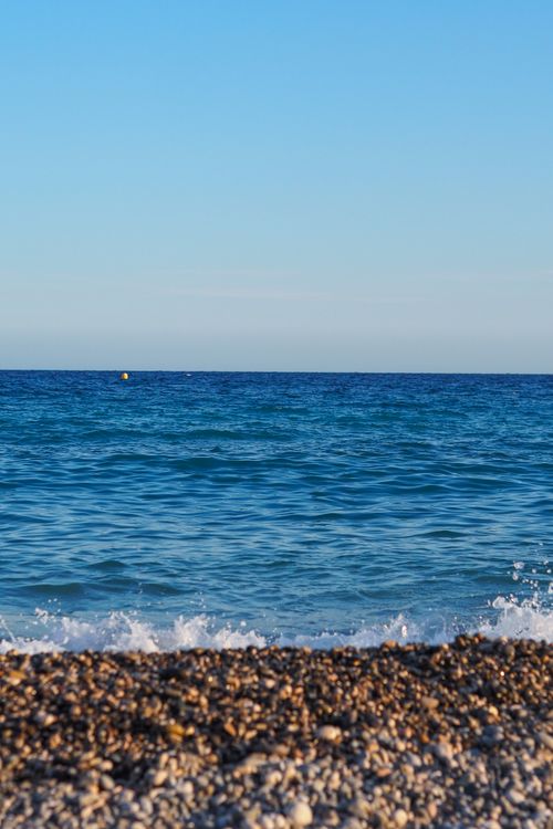 Free Serene pebble beach with gentle ocean waves under a clear blue sky. Stock Photo