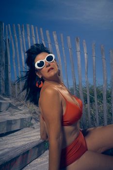 Stylish woman in red swimsuit and sunglasses relaxing by a wooden beach fence on a sunny day.