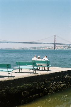 Three people enjoy a peaceful view of the Ponte 25 de Abril in Lisbon from a bench by the river.