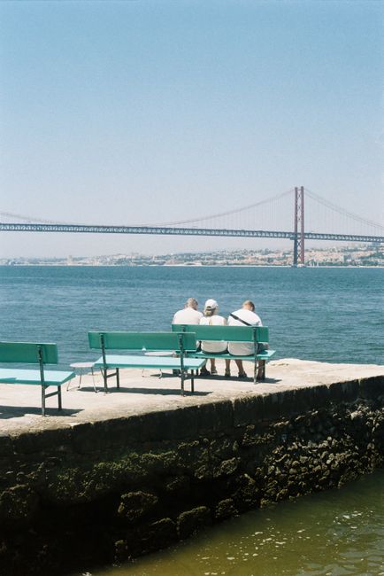 Three people enjoy a peaceful view of the Ponte 25 de Abril in Lisbon from a bench by the river.