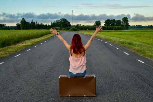 Red-haired woman sitting on a suitcase in the middle of a country road, arms raised in freedom.
