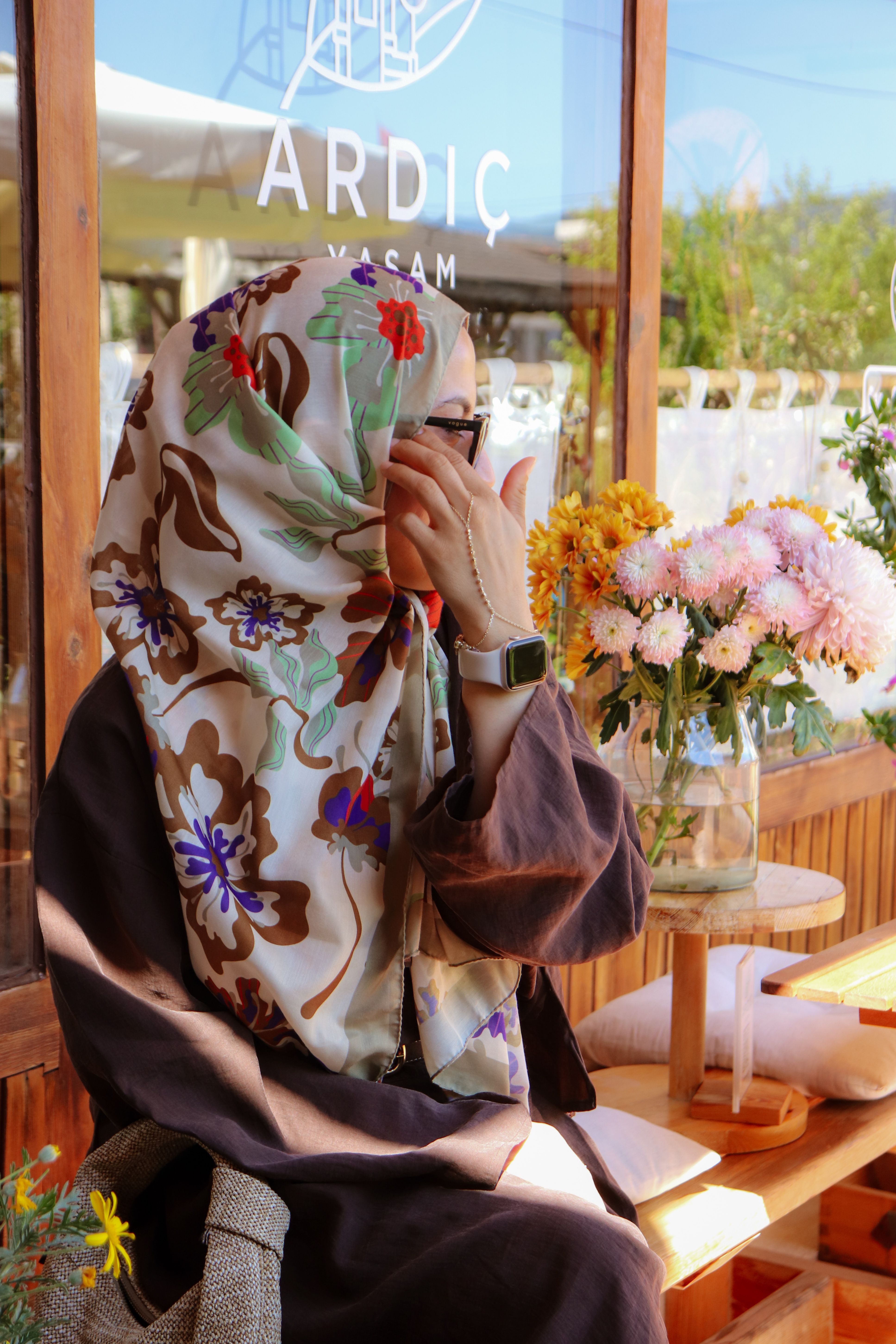 A woman in a floral hijab seated at an outdoor café, surrounded by vibrant flowers.
