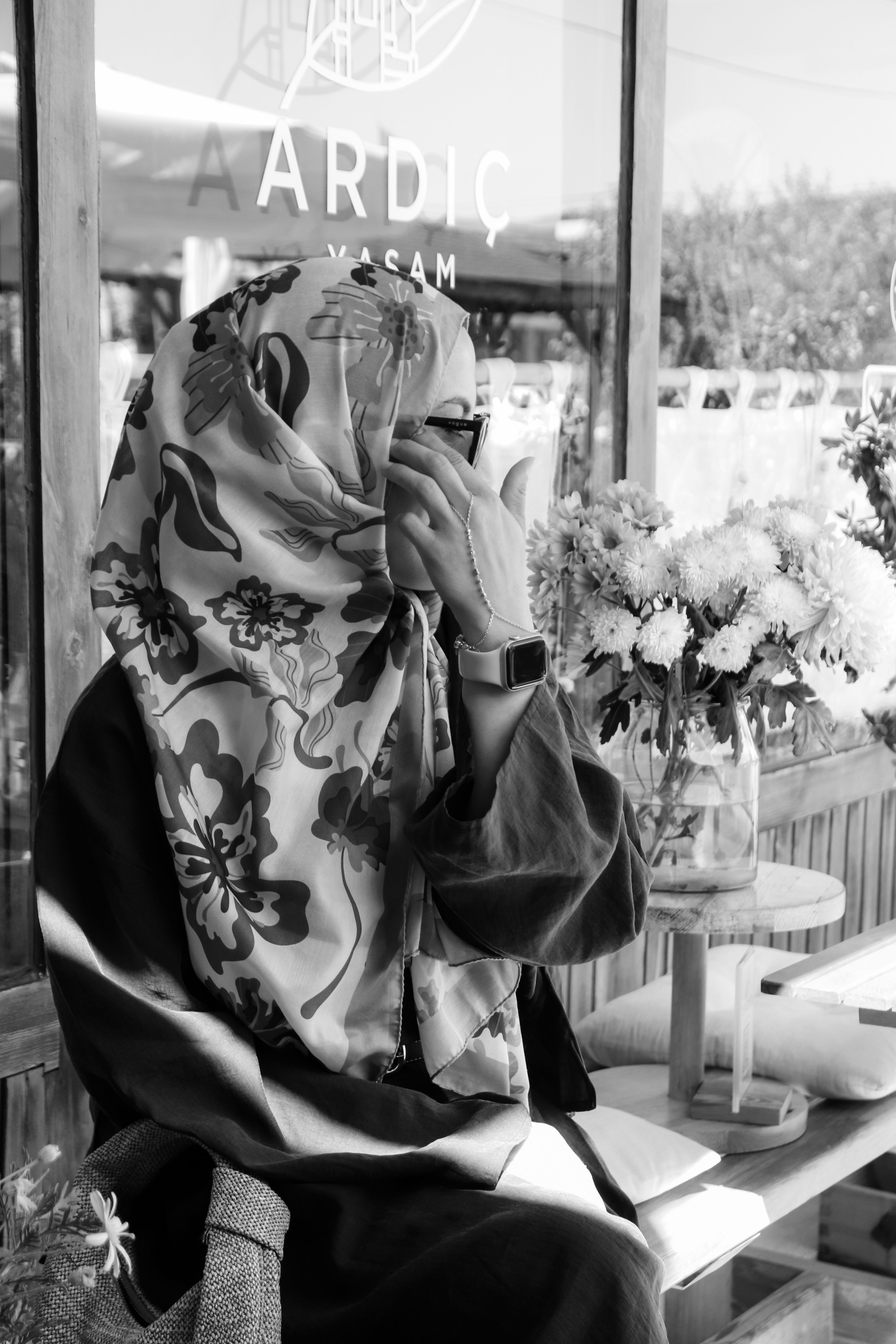 Woman in sunglasses and floral hijab sitting in cafe, monochrome style.