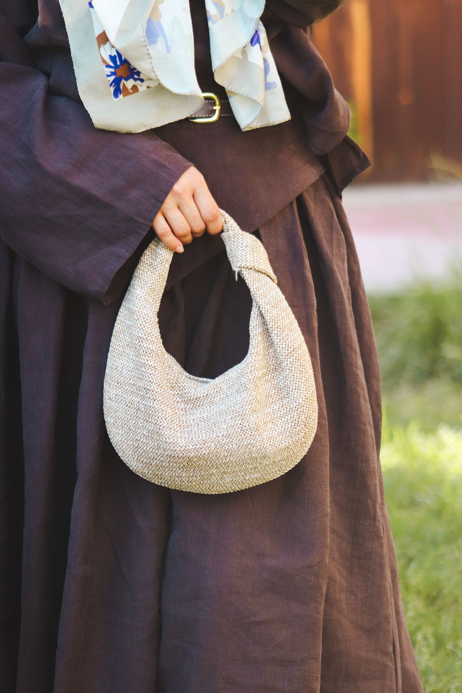 Close-up of a model in a brown dress holding a woven handbag with a floral scarf.