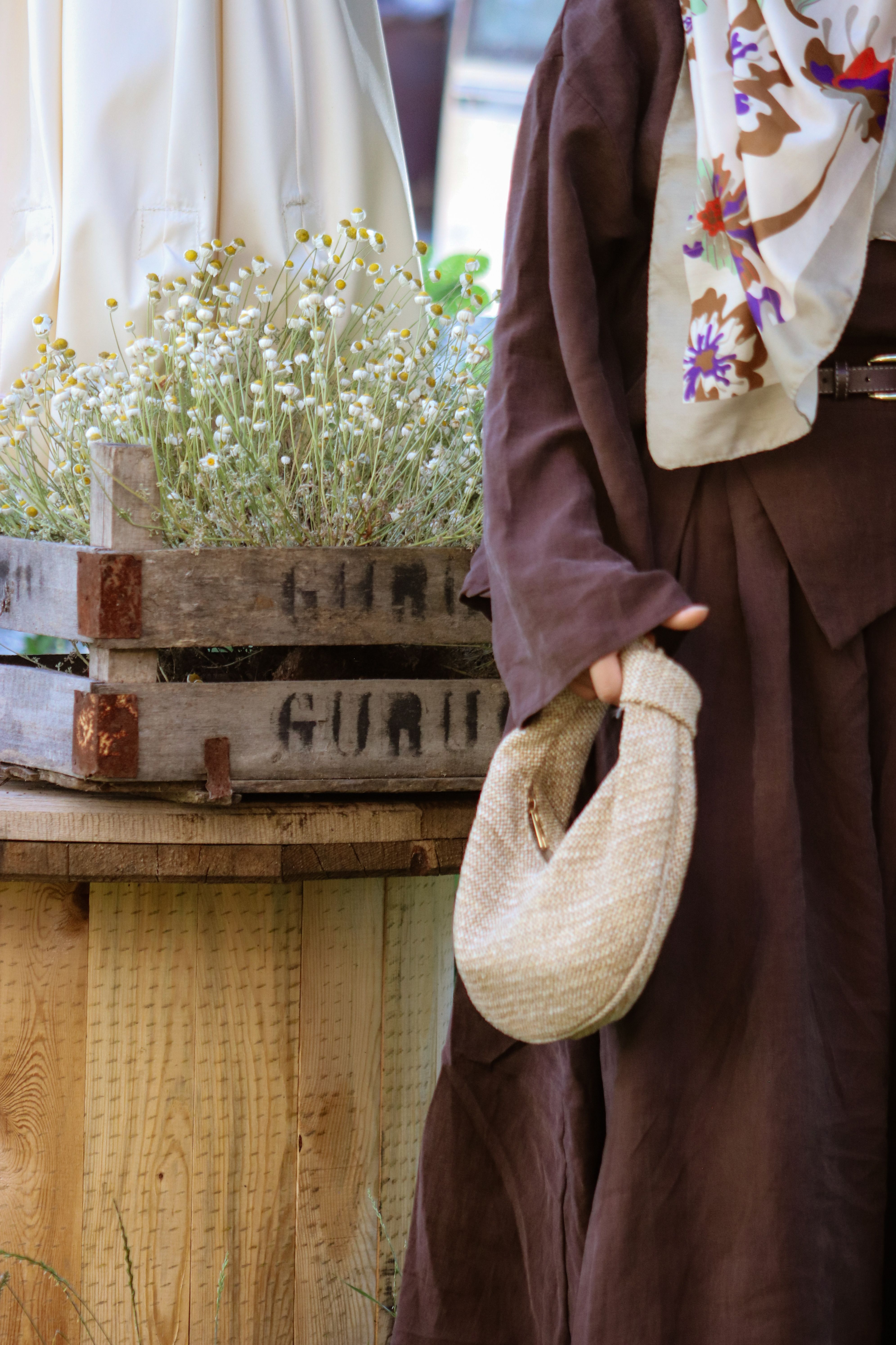 A woman in a brown outfit holds a wicker bag near a rustic flower arrangement outdoors.