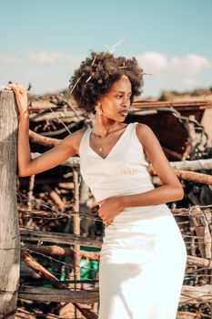 Elegant African woman in a white dress poses outdoors in a rustic setting, showcasing natural beauty and fashion.