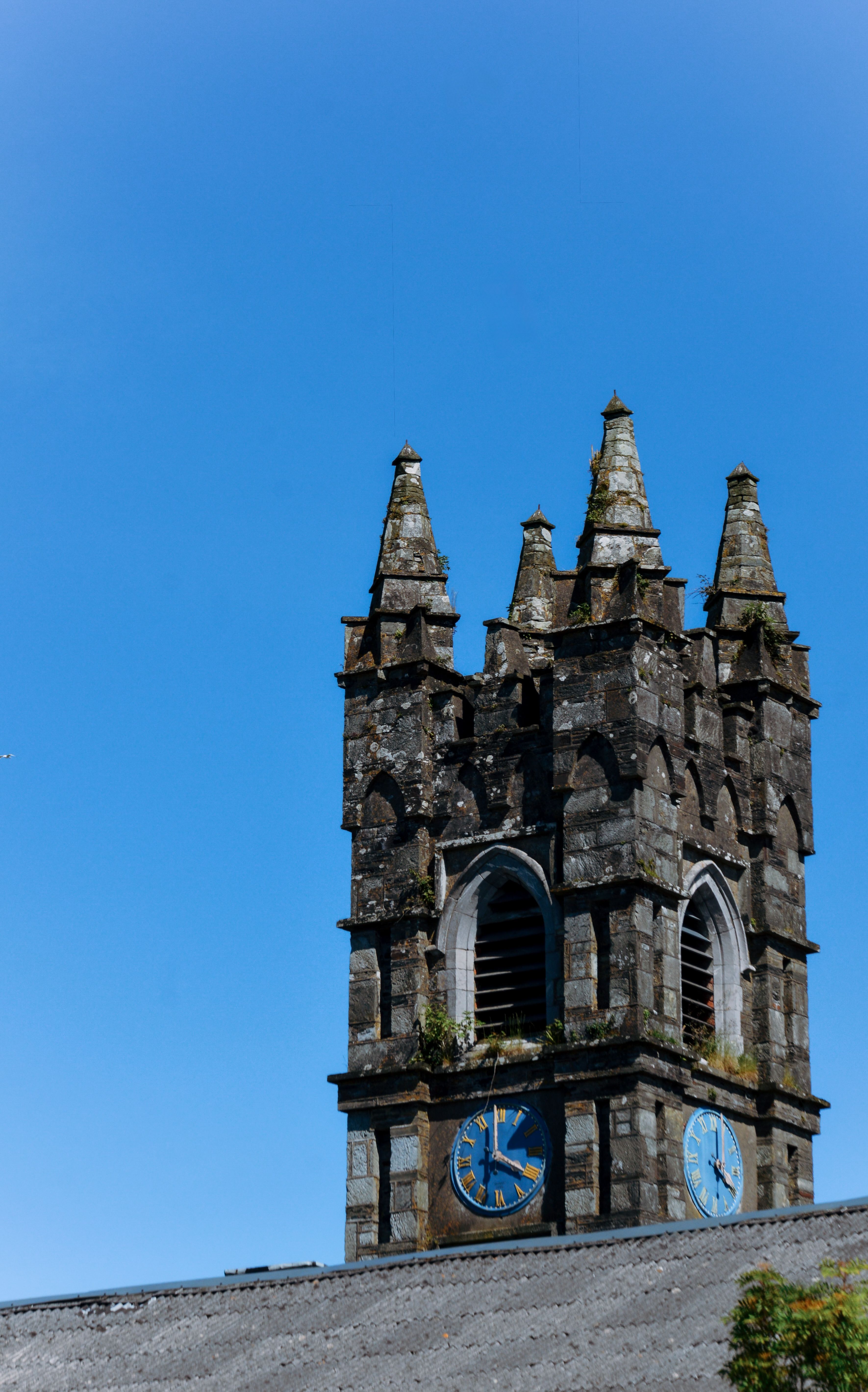 A historic stone church tower with blue clock faces in Bantry, Ireland under clear skies.