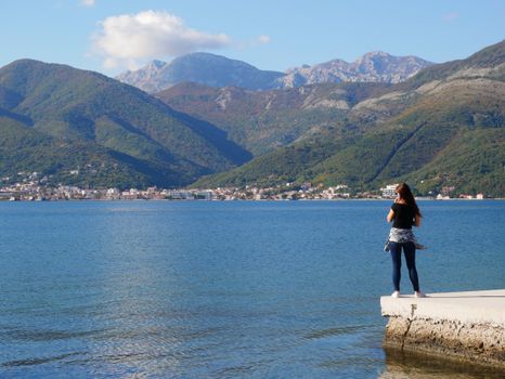 A woman stands on a pier, admiring the scenic bay and mountains in Tivat, Montenegro.