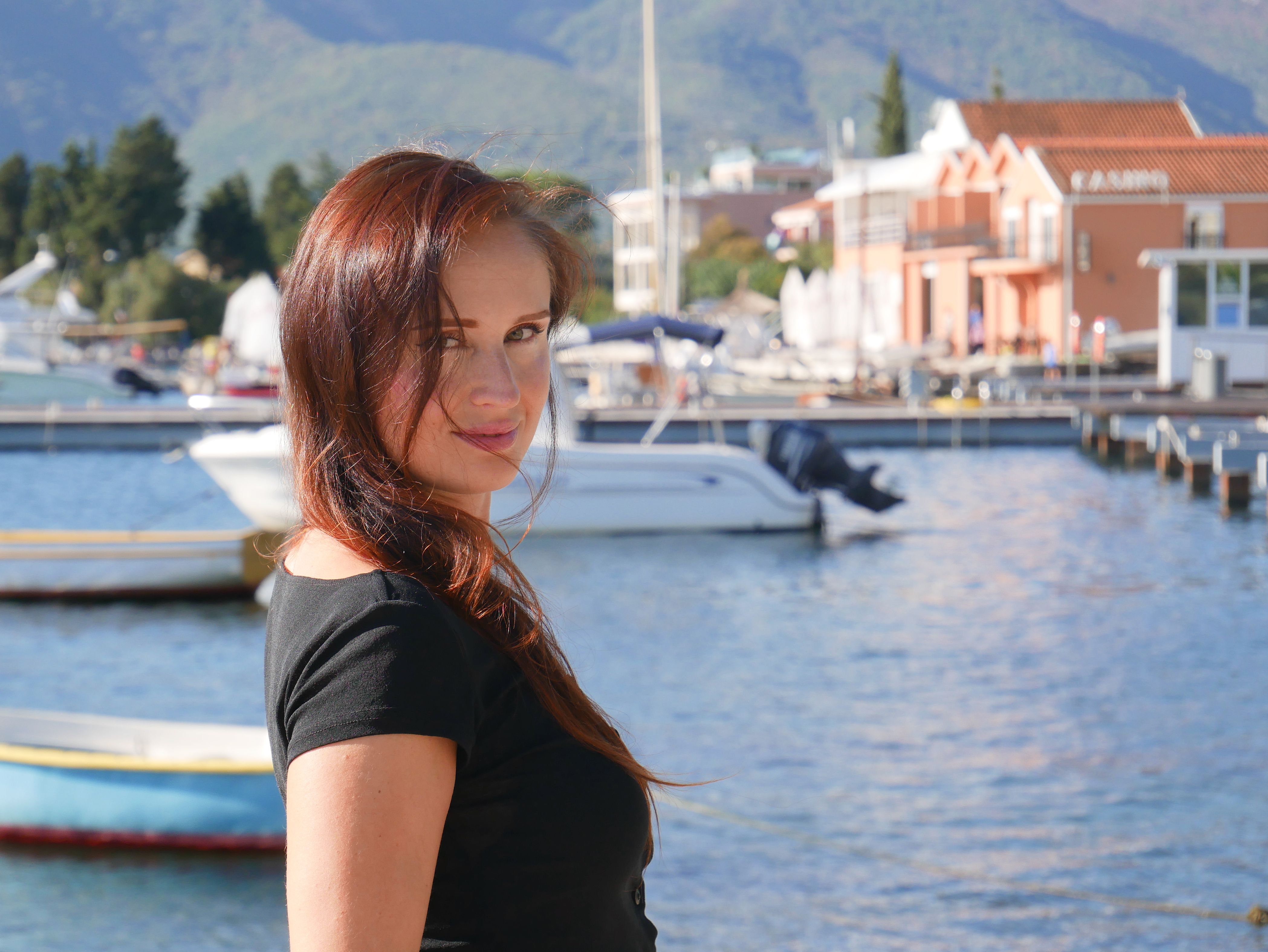 Portrait of a woman smiling at Tivat marina with boats and mountains in the background.