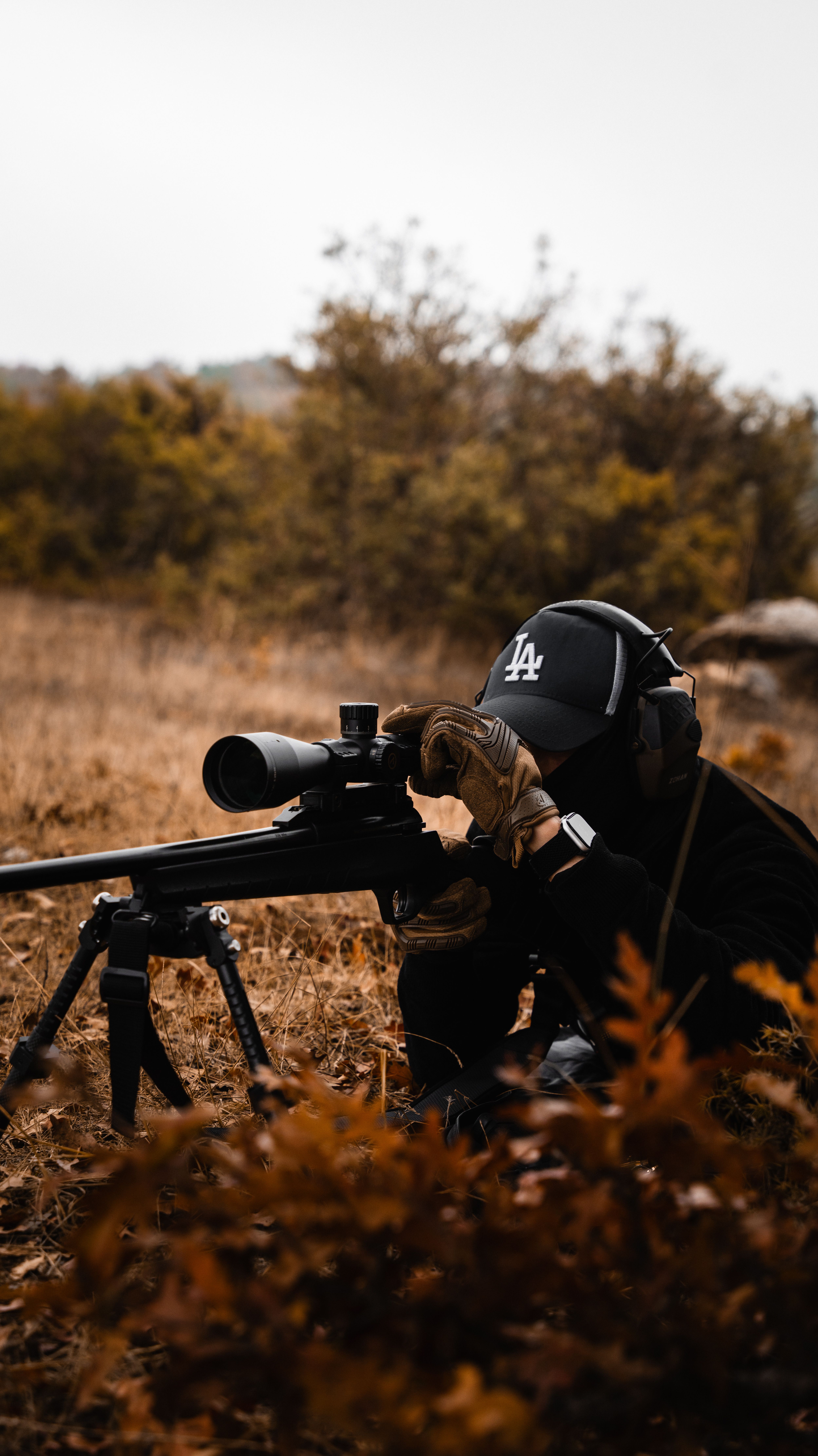 A sniper in tactical gear aiming a rifle outdoors, surrounded by autumn foliage.