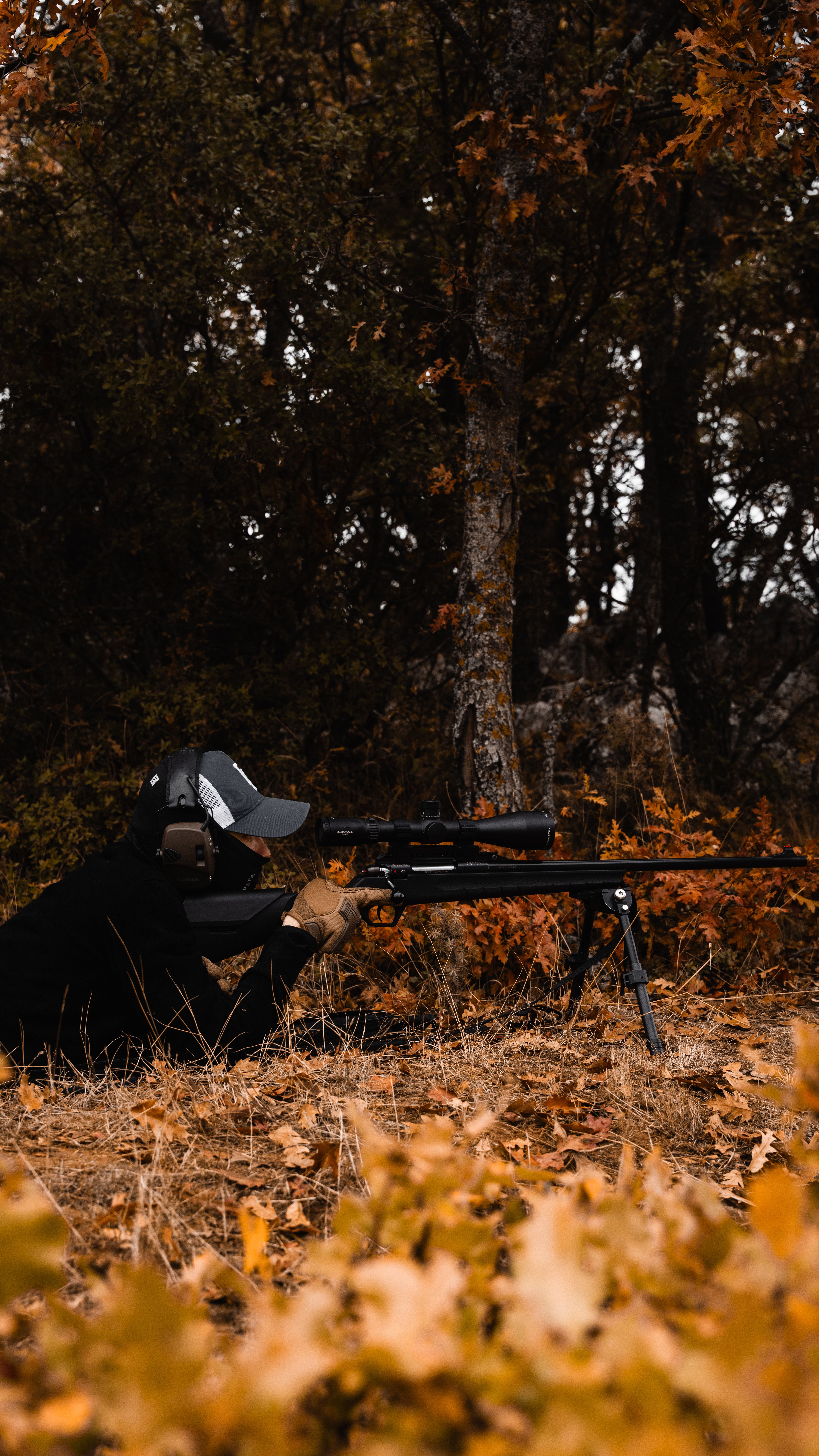 A camouflaged person aiming a rifle in the autumn forest, surrounded by brown foliage.