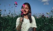 一位身着白色礼服的年轻民族女性，手持花茎，在晴天的蓝天下，面对开花的种植园站立