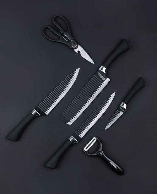 Top view of various kitchen utensils including knives scissors and vegetable peeler on black background